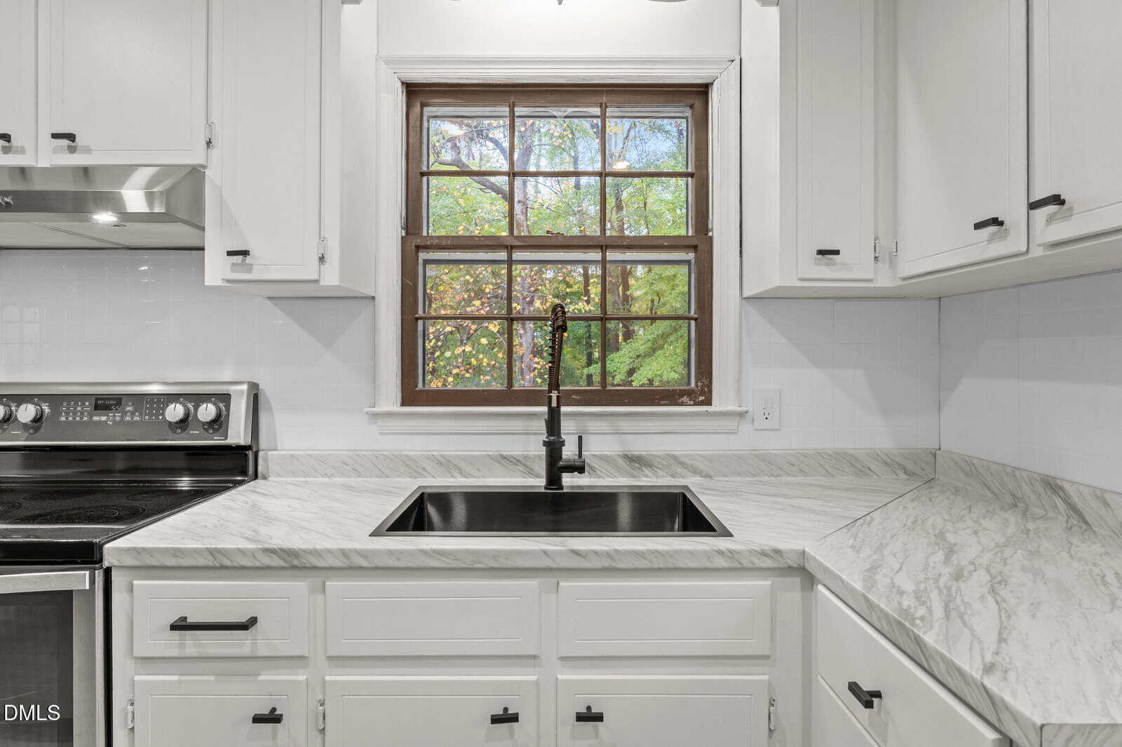 A kitchen with white cabinets, a marble-patterned countertop, a black sink with a black faucet, an electric stove, and a window above the sink showing green trees outside.