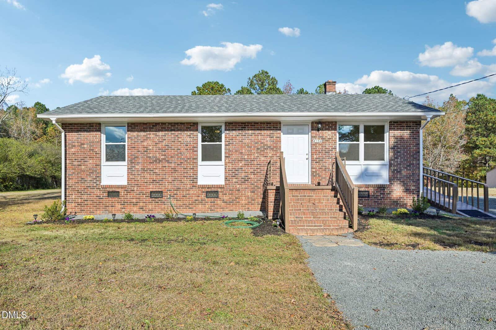 A single-story brick house in Oxford at 4158 Shock Overton Road features a gray shingle roof, white door, three front windows, brick steps, a gravel driveway, and a wheelchair-accessible ramp. The grassy yard is dotted with small plants.