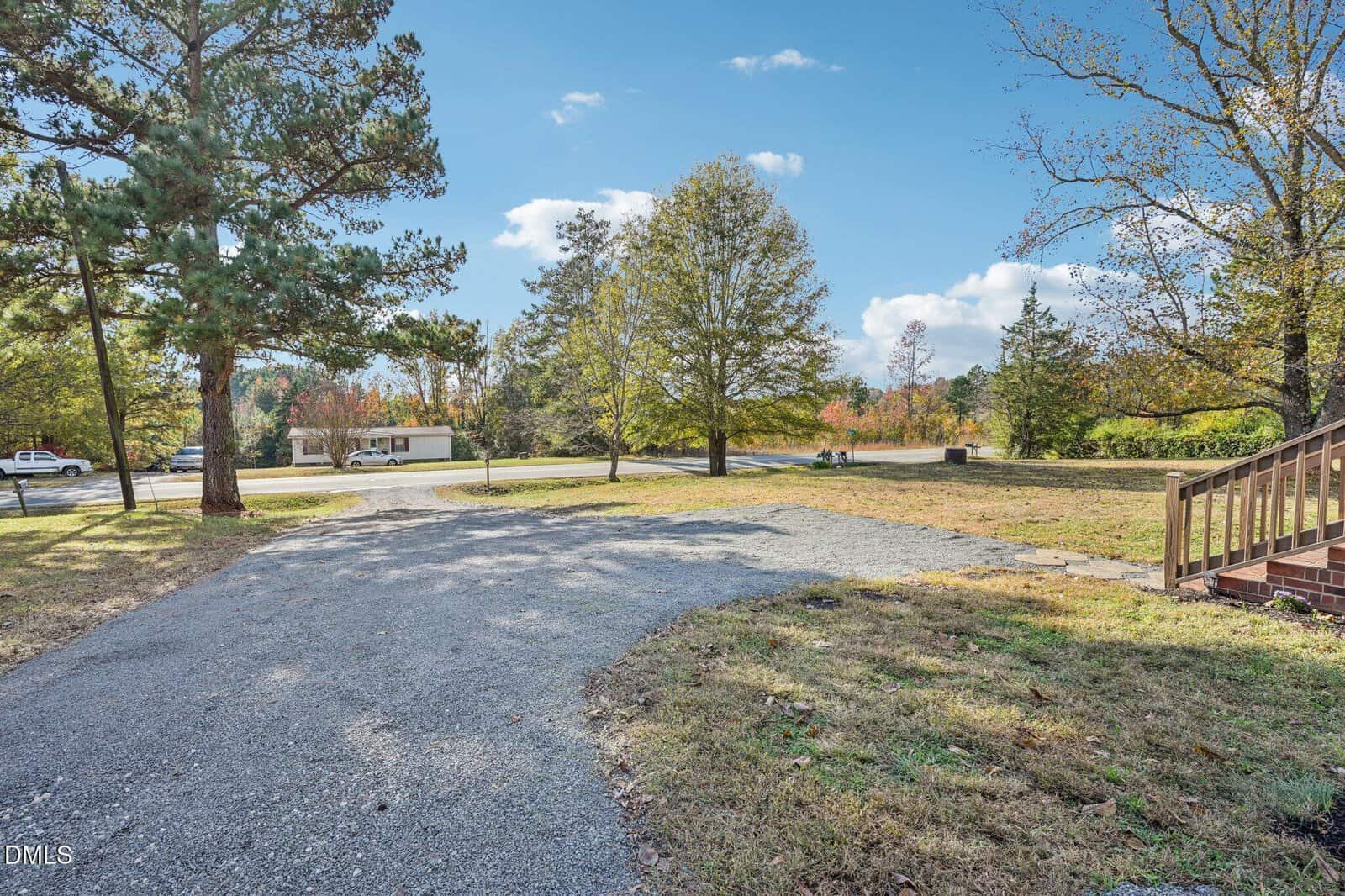A gravel driveway curves toward a residential street lined with trees. A patch of grass borders the drive, and wooden steps lead up to a porch on the right. A white car and a house are visible across the street.