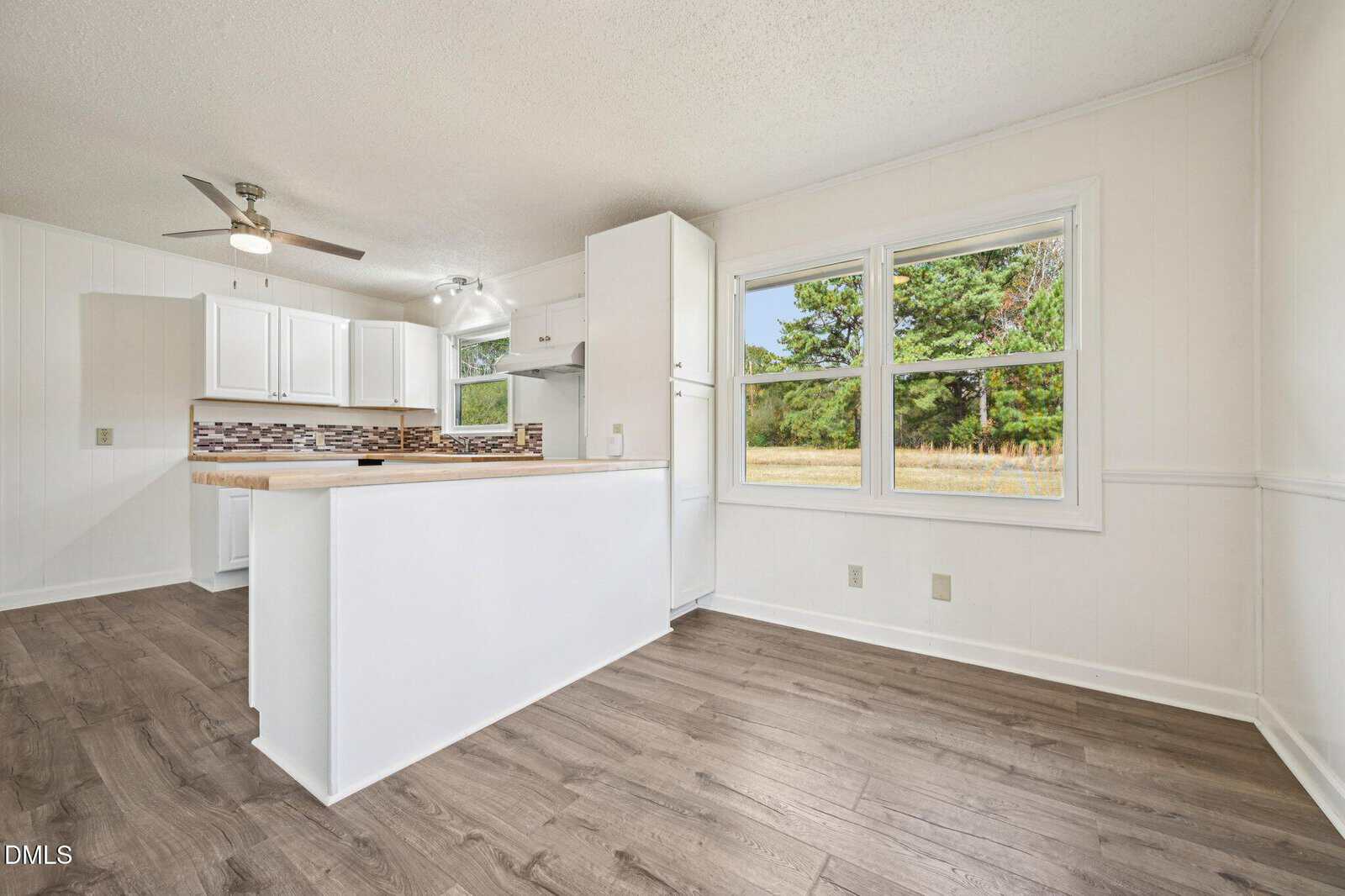 Bright kitchen and dining area with white walls, wood-style flooring, white cabinets, and a kitchen island. Large windows provide a view of trees and a grassy yard outside. Ceiling fans and modern lighting are visible.