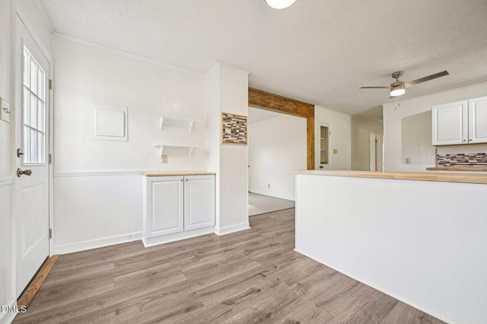 Bright kitchen area with white cabinets, wood countertops, light wood flooring, white walls, open shelving, a ceiling fan, and a doorway leading to another room.