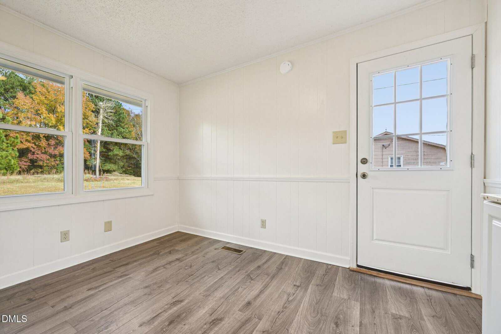 A small, empty room with white paneled walls, wood-look flooring, two windows showing trees outside, and a white door with a window grid and metal handle.