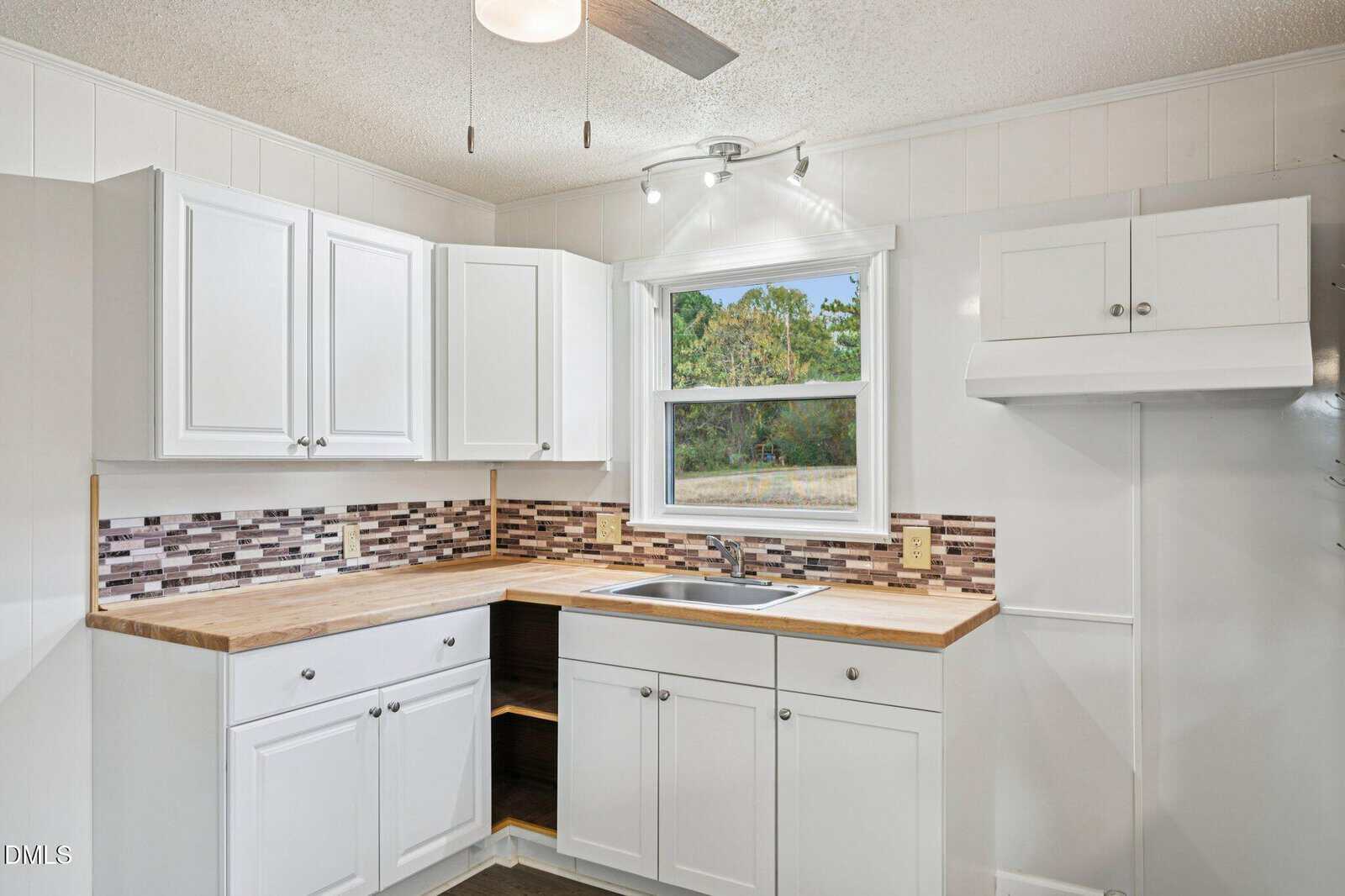 A small kitchen with white cabinets, wood countertops, mosaic tile backsplash, a stainless steel sink below a window, open shelving, and a ceiling light with a fan. A view of trees is visible through the window.