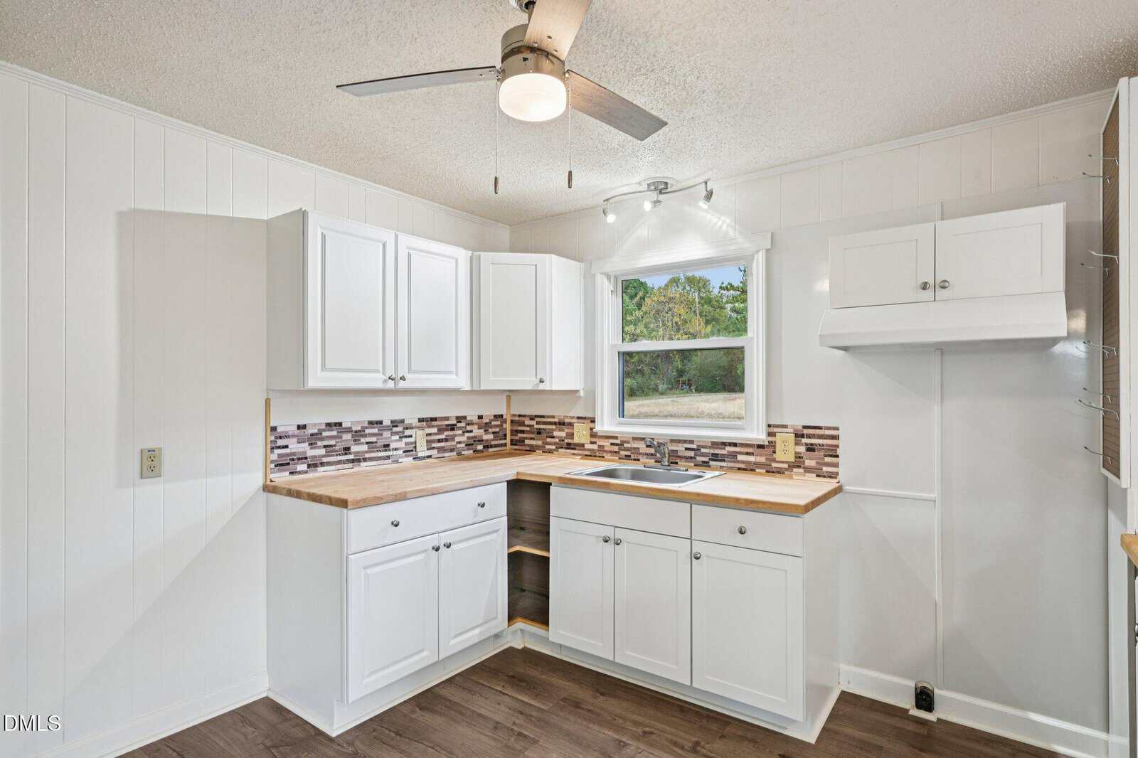 A small kitchen with white cabinets, wood countertops, a mosaic tile backsplash, a stainless steel sink under a window, and dark wood flooring. A ceiling fan with a light is mounted on the white ceiling.
