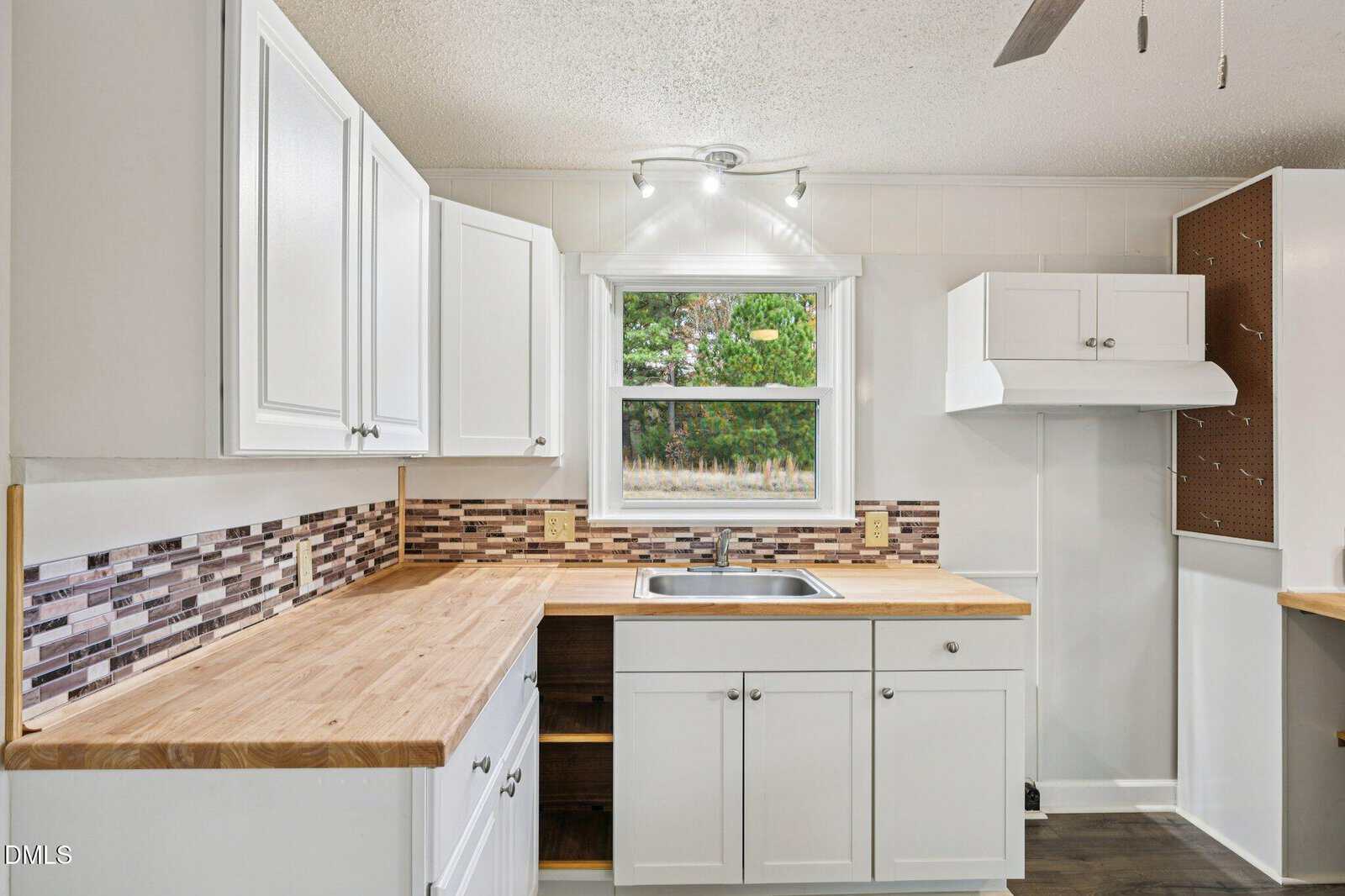 A kitchen with white cabinets, wood countertops, a stainless steel sink under a window, and a tile mosaic backsplash. The walls are white, and there is a ceiling fan and pegboard on the right side.