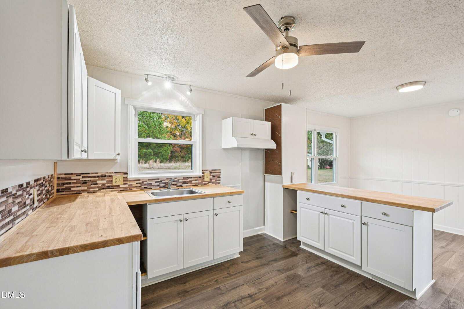 Bright kitchen with white cabinets, wood countertops, mosaic tile backsplash, stainless steel sink, ceiling fan, and large window. A peninsula counter provides extra space. The floor is dark wood.