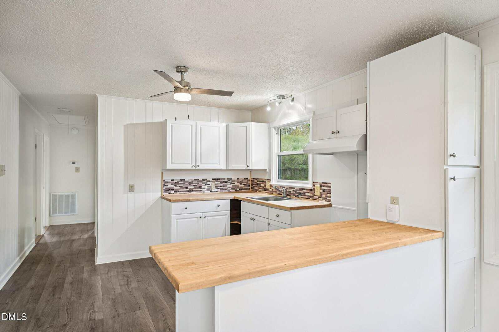 Bright kitchen with white cabinets, wood countertops, tile backsplash, stainless steel ceiling fan, and window above the sink. Light wood flooring and white walls create a clean, modern look.