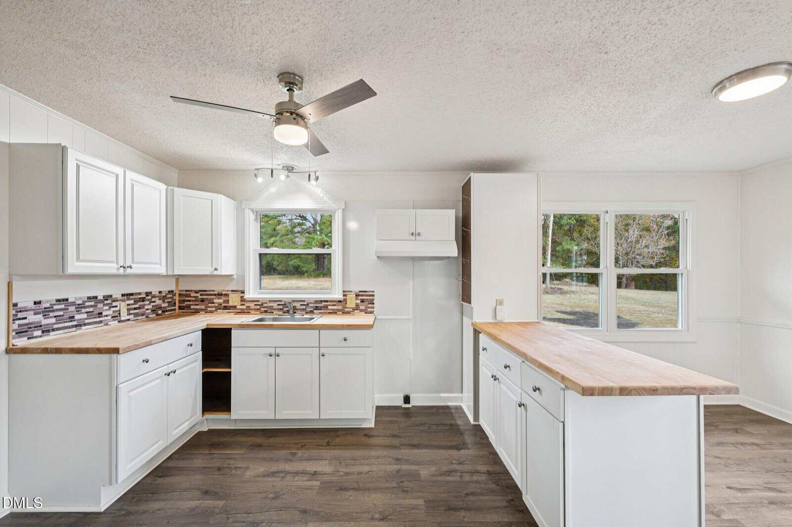 Modern kitchen with white cabinets, wood countertops, a tile backsplash, stainless steel ceiling fan, and two windows providing outdoor views. The floor is dark wood, and the space is well-lit and open.