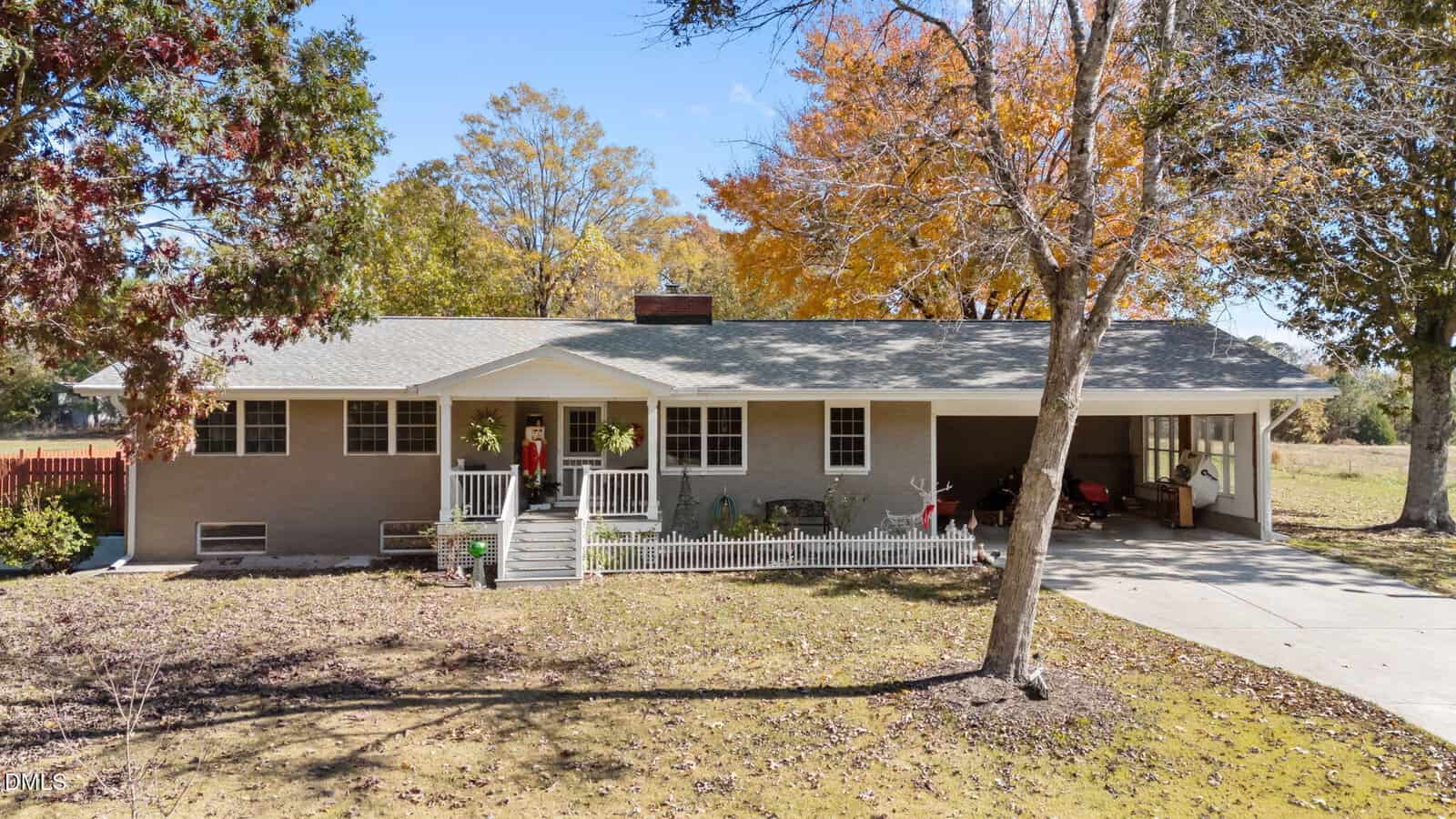 Single-story house with gray siding, front porch, and white railing located at 444 Barnette Road in Louisburg. A carport is attached to the right side. Trees with autumn leaves surround the house, and the front yard has sparse grass and scattered shade.