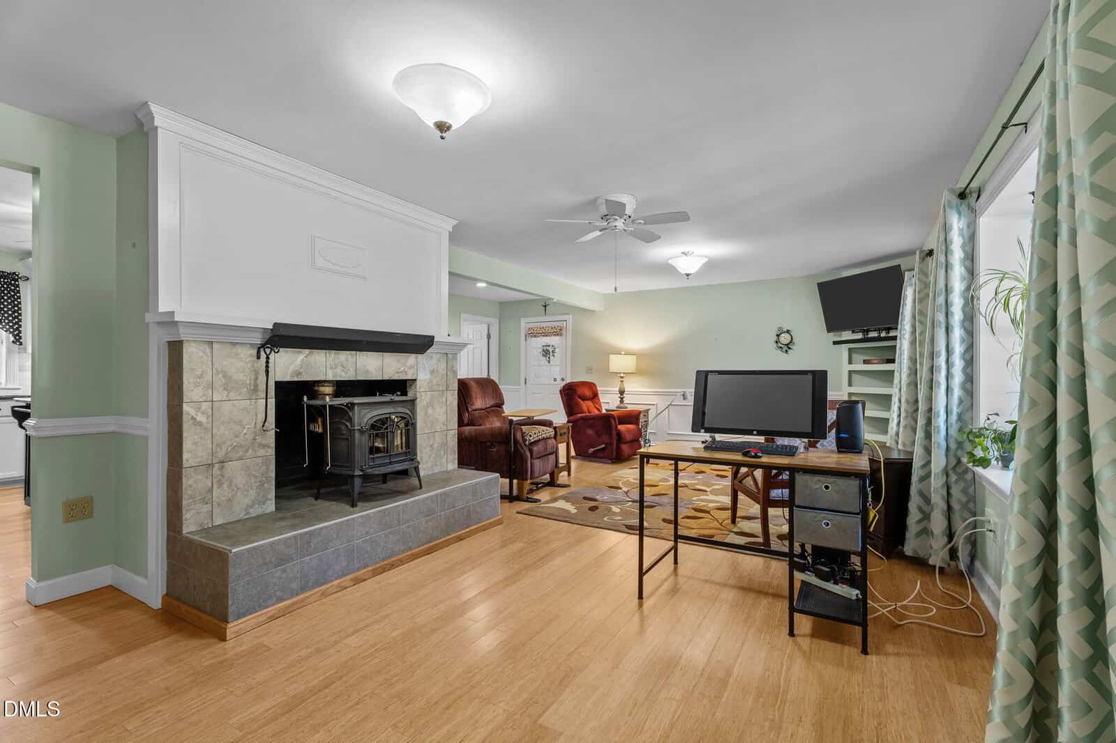 Open living area with a wood stove on a tiled platform, two recliners, a desk with a computer, a wall-mounted TV, bamboo flooring, and patterned curtains by a large window.