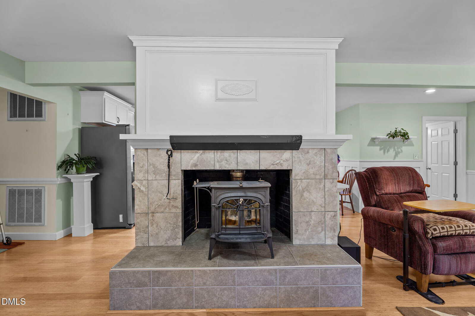 A wood-burning stove sits in a tiled fireplace with a white mantel, in a living room with hardwood floors, a red armchair, and a view of a kitchen area with green walls and white cabinetry.