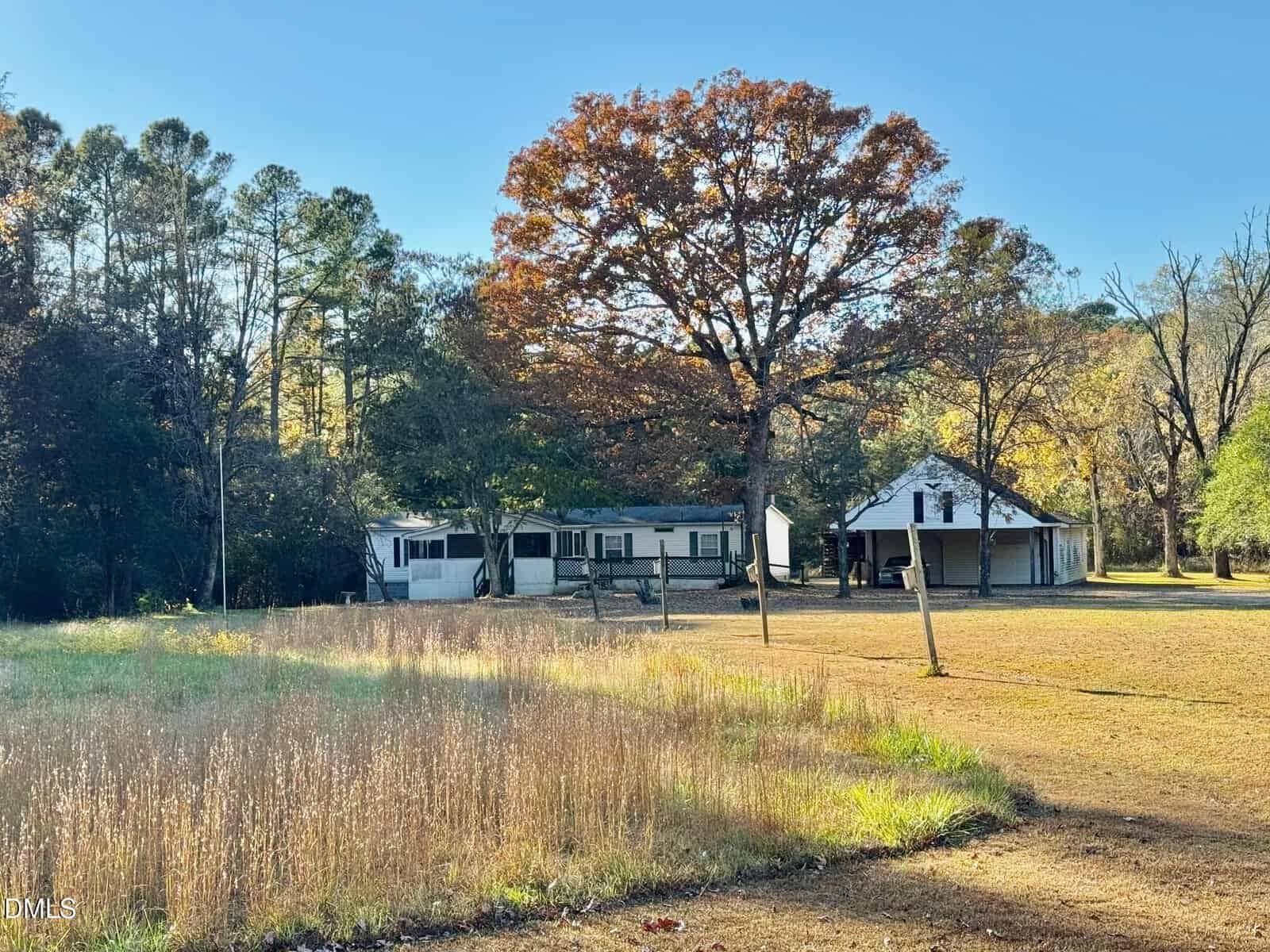 A white house and a separate garage or shed sit in a large, grassy yard with a mix of tall and short grass. Trees with autumn foliage surround the property under a clear blue sky.