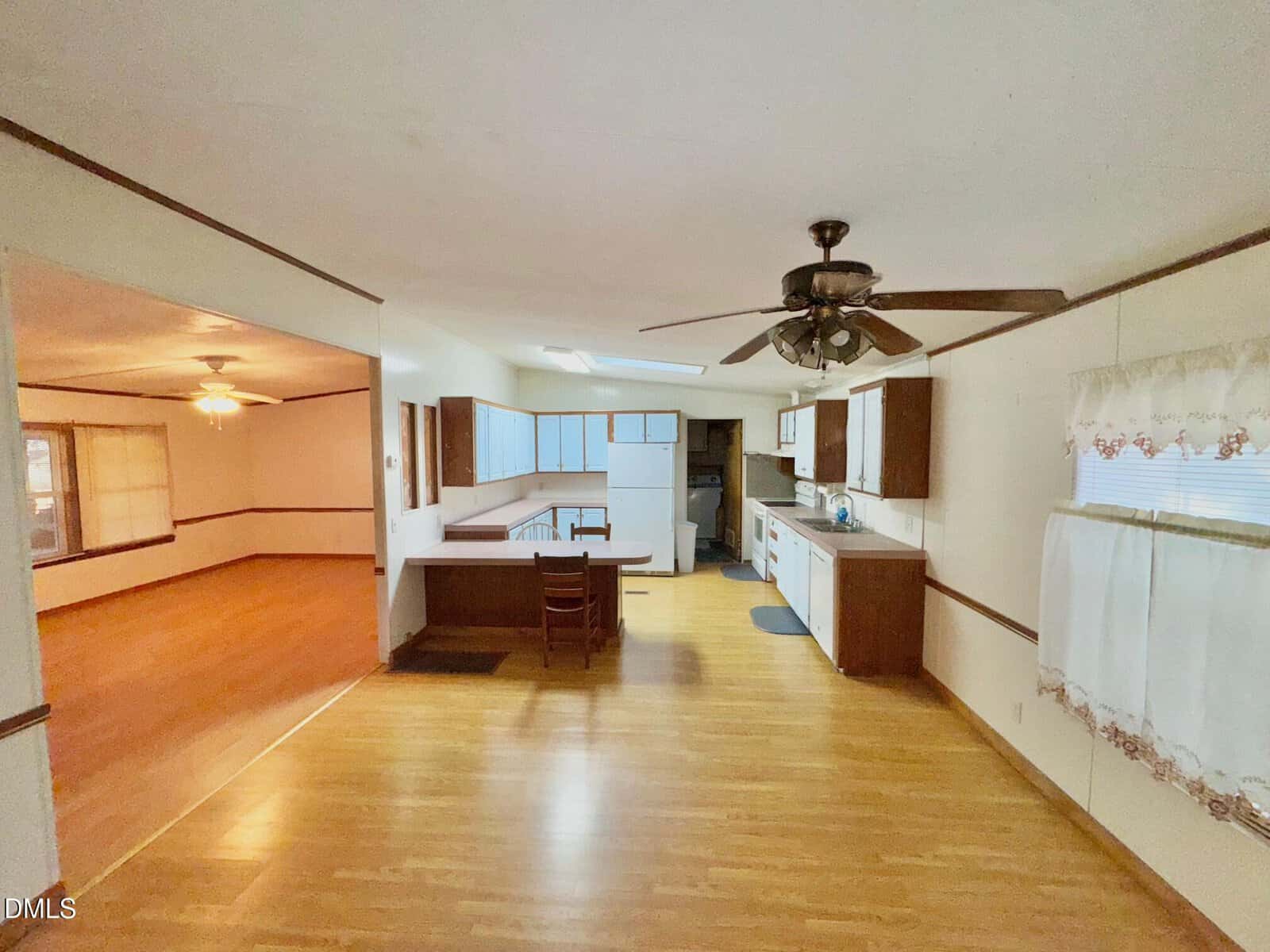 Spacious kitchen with wood floors, white countertops, brown cabinets, ceiling fan, and dining area. Adjacent room with wood floors and another ceiling fan is visible through a wide doorway. Large windows provide natural light.