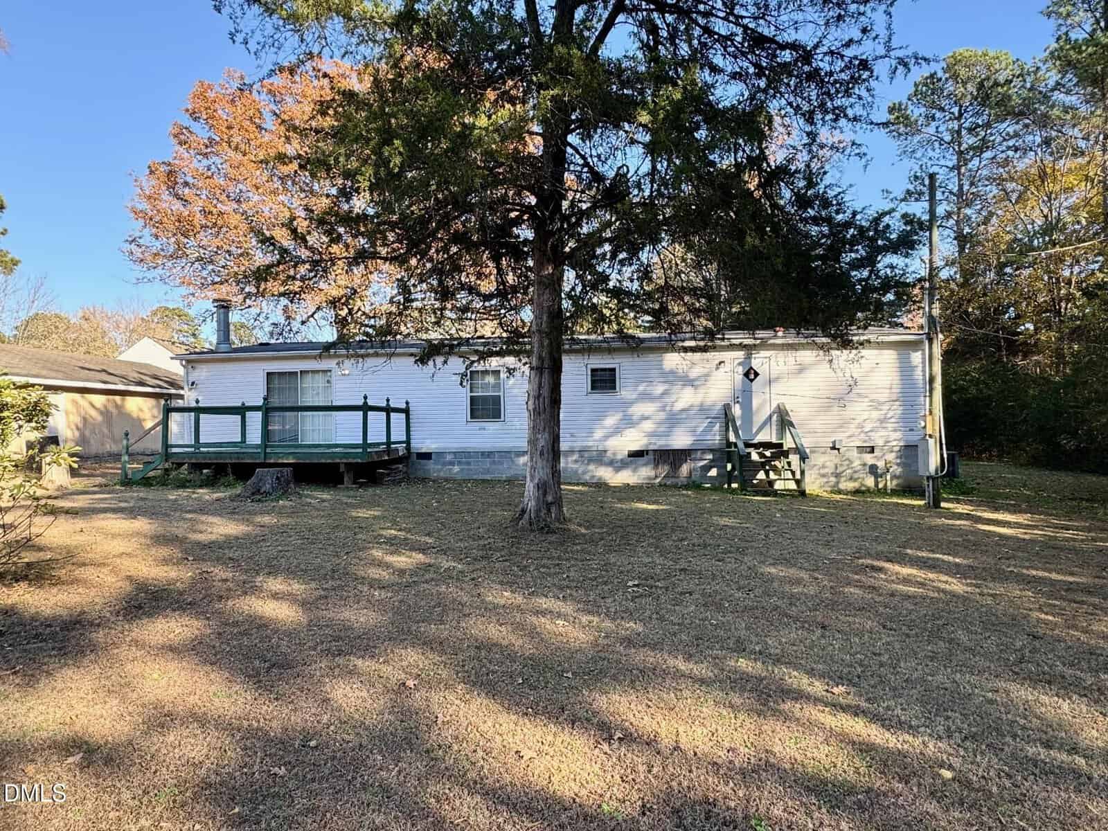 Single-story white house with green railings, small porch steps, and a wooden deck. A large tree stands in the foreground. The yard is grassy with some patchy areas and wooded surroundings. Sky is clear and blue.
