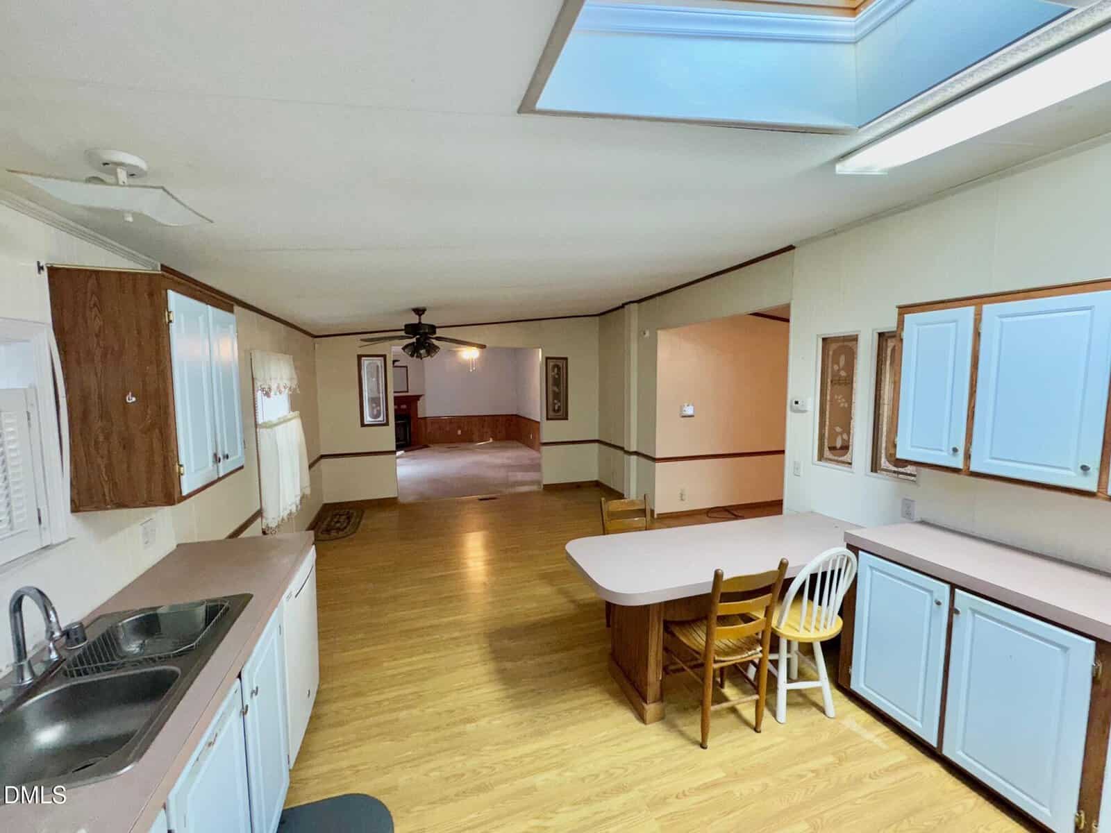 A spacious open kitchen with white cabinets, wooden accents, a sink, and a small dining area with two chairs. The space connects to a larger living area with wood flooring and ceiling fans. Skylight and overhead lights are visible.