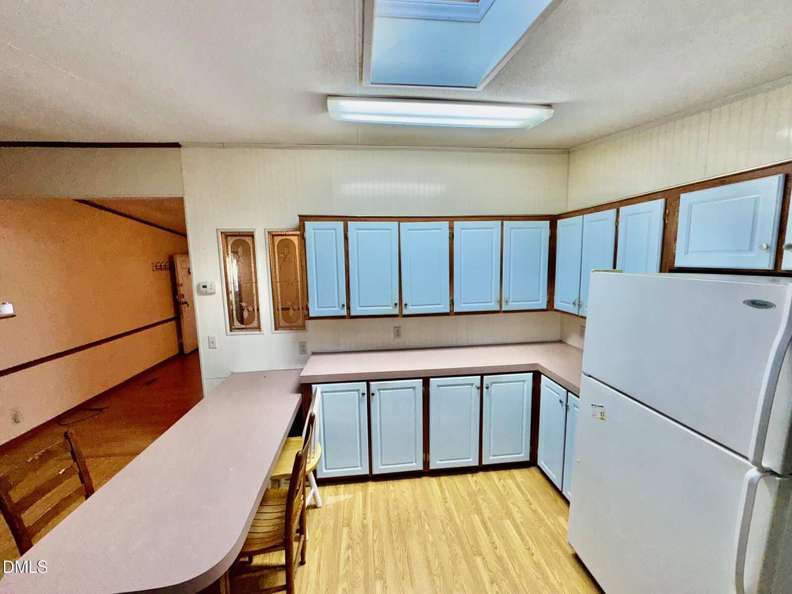 A kitchen with light blue cabinets, white countertops, a white refrigerator, and wood flooring. There are two chairs at a counter, a fluorescent ceiling light, and a skylight above. The walls are off-white.