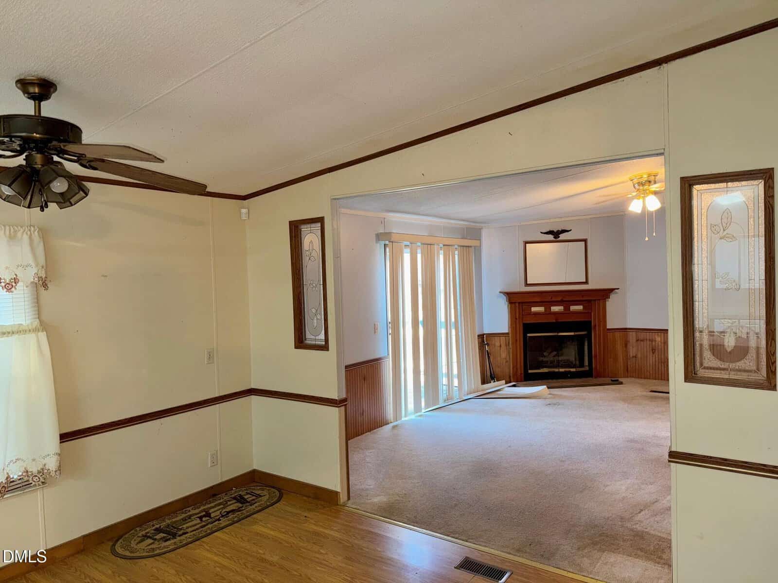 Interior of a home showing two rooms: one with wood flooring, ceiling fan, and window; the other with carpet, a fireplace, ceiling fan, large mirror, and sliding glass doors with vertical blinds.