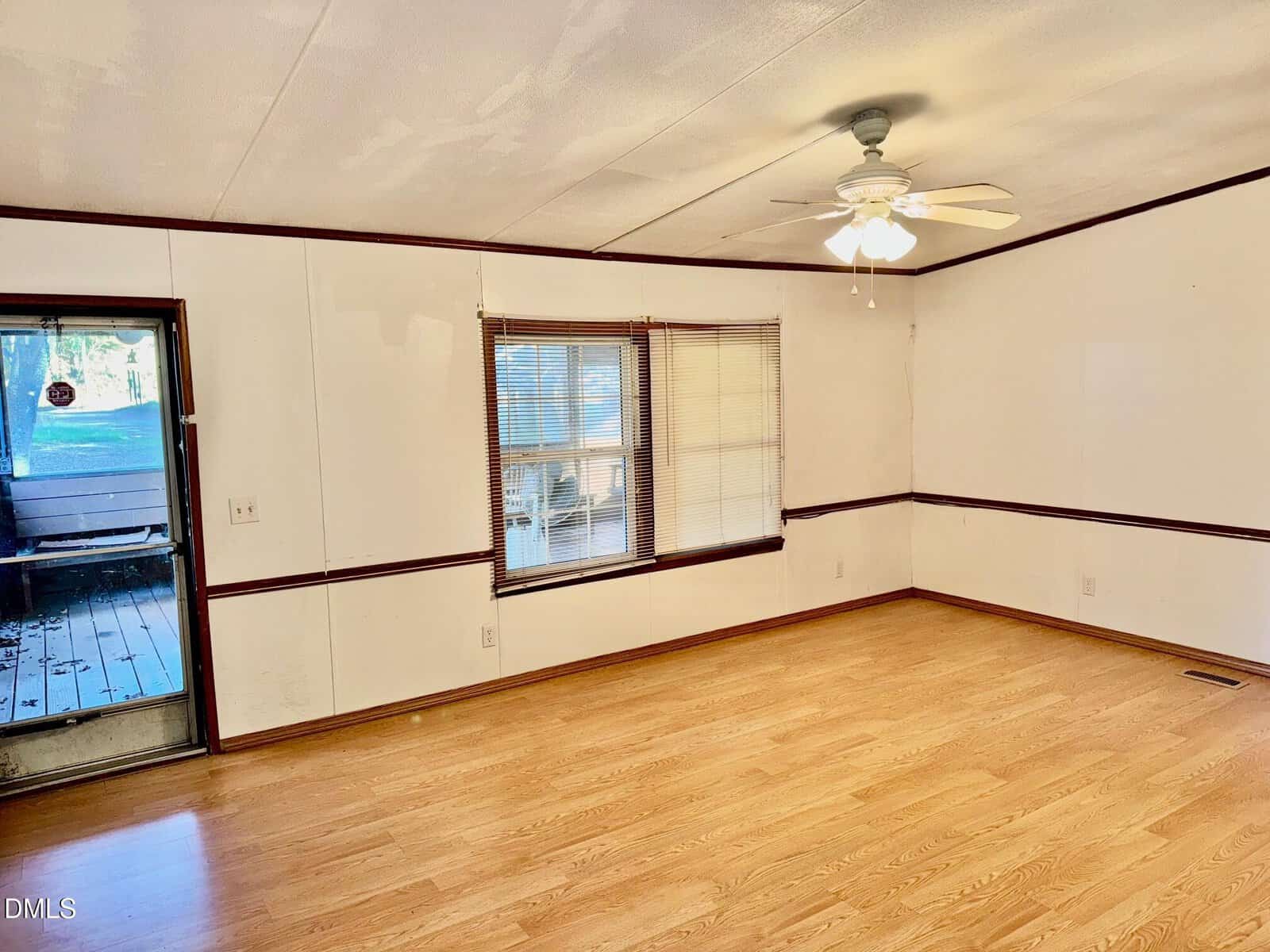 An empty room with light wood flooring, white walls with dark trim, a ceiling fan with lights, two windows with blinds, and a door leading to a covered porch.
