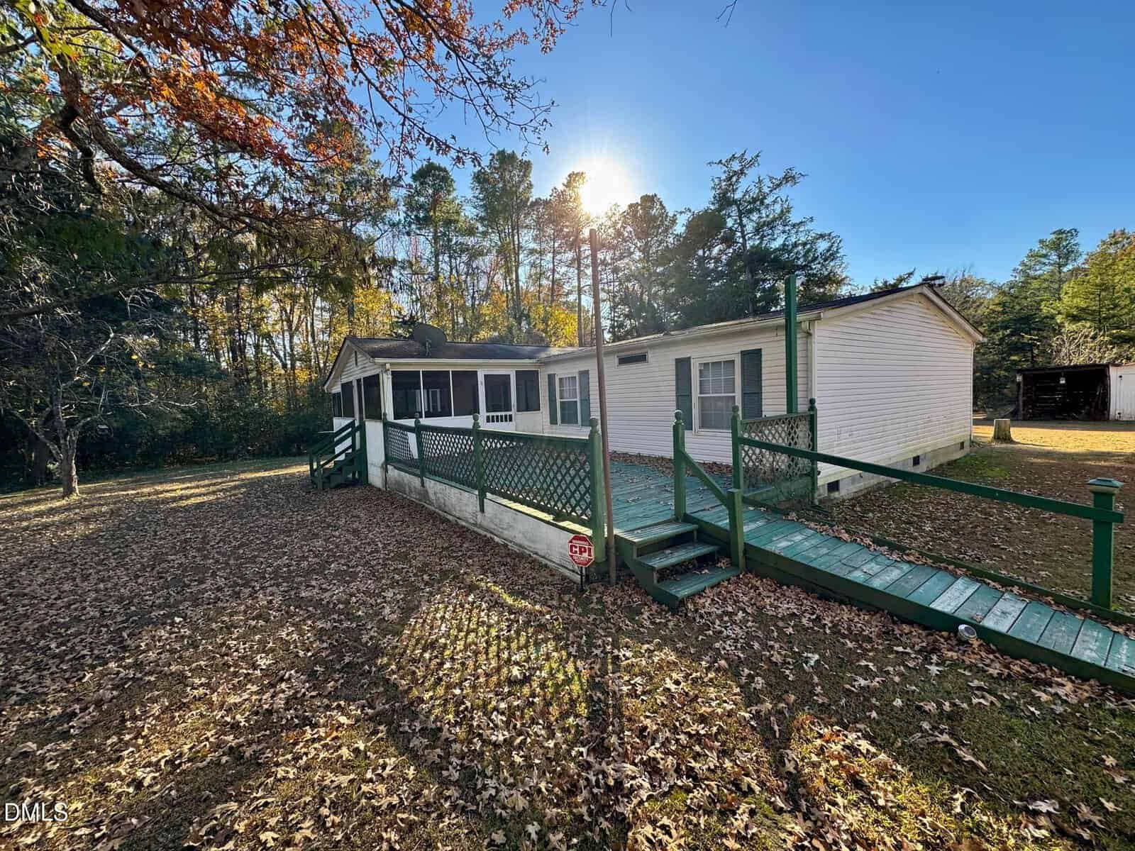 Single-story white house with green trim and a green ramp leading to the entrance. The yard is covered with fallen leaves, surrounded by trees, under a clear blue sky with sunlight filtering through.