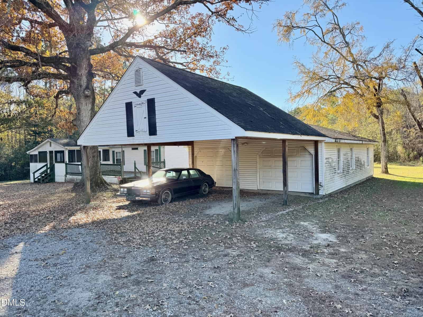 A black car is parked under a white open carport attached to a house with a gabled roof. Trees with autumn leaves surround the area, and another small building is visible in the background.