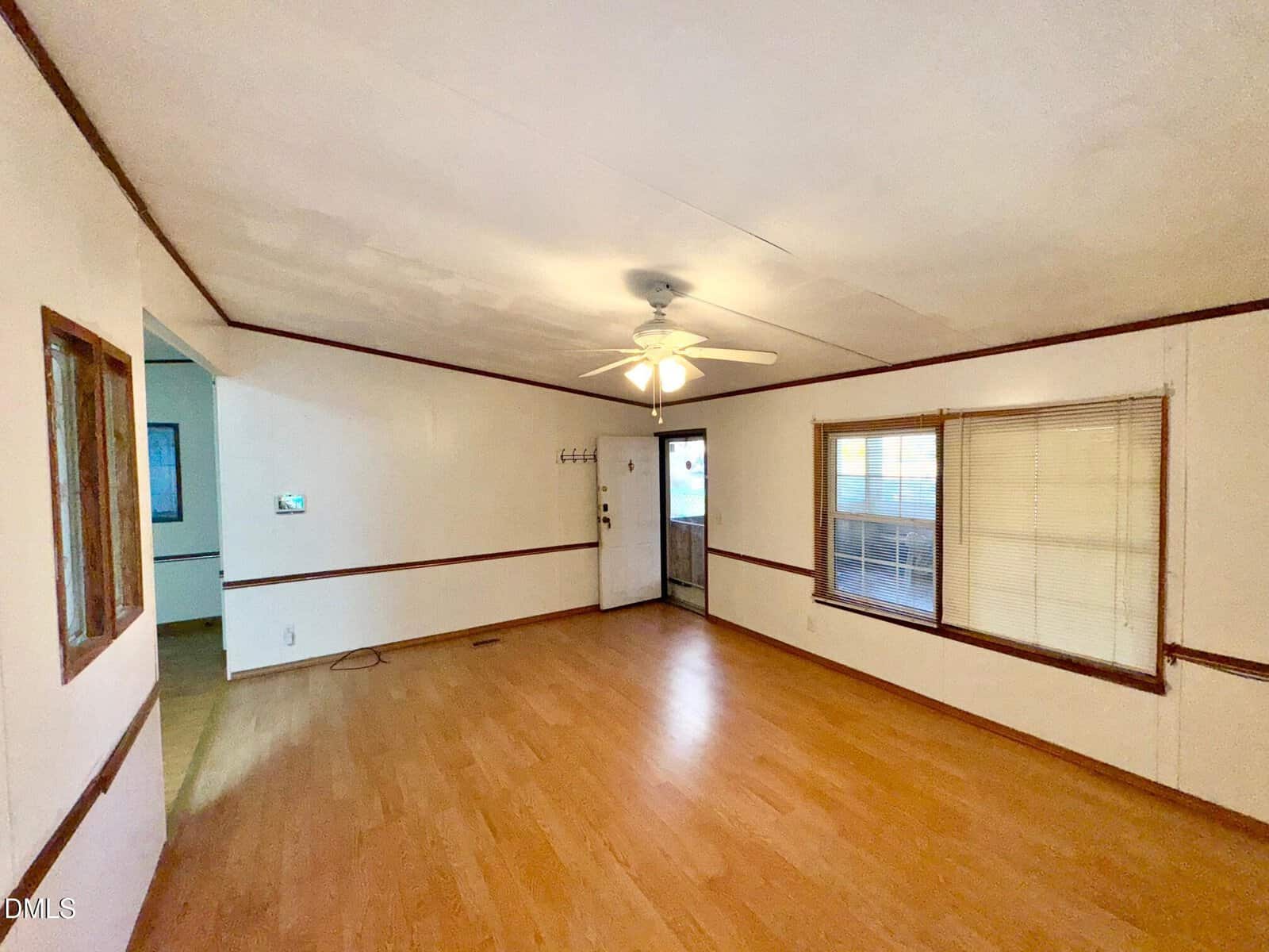 Empty living room with wood laminate flooring, white walls, brown trim, a ceiling fan with light, several windows, and a door leading outside. Room is unfurnished and well-lit by natural light.