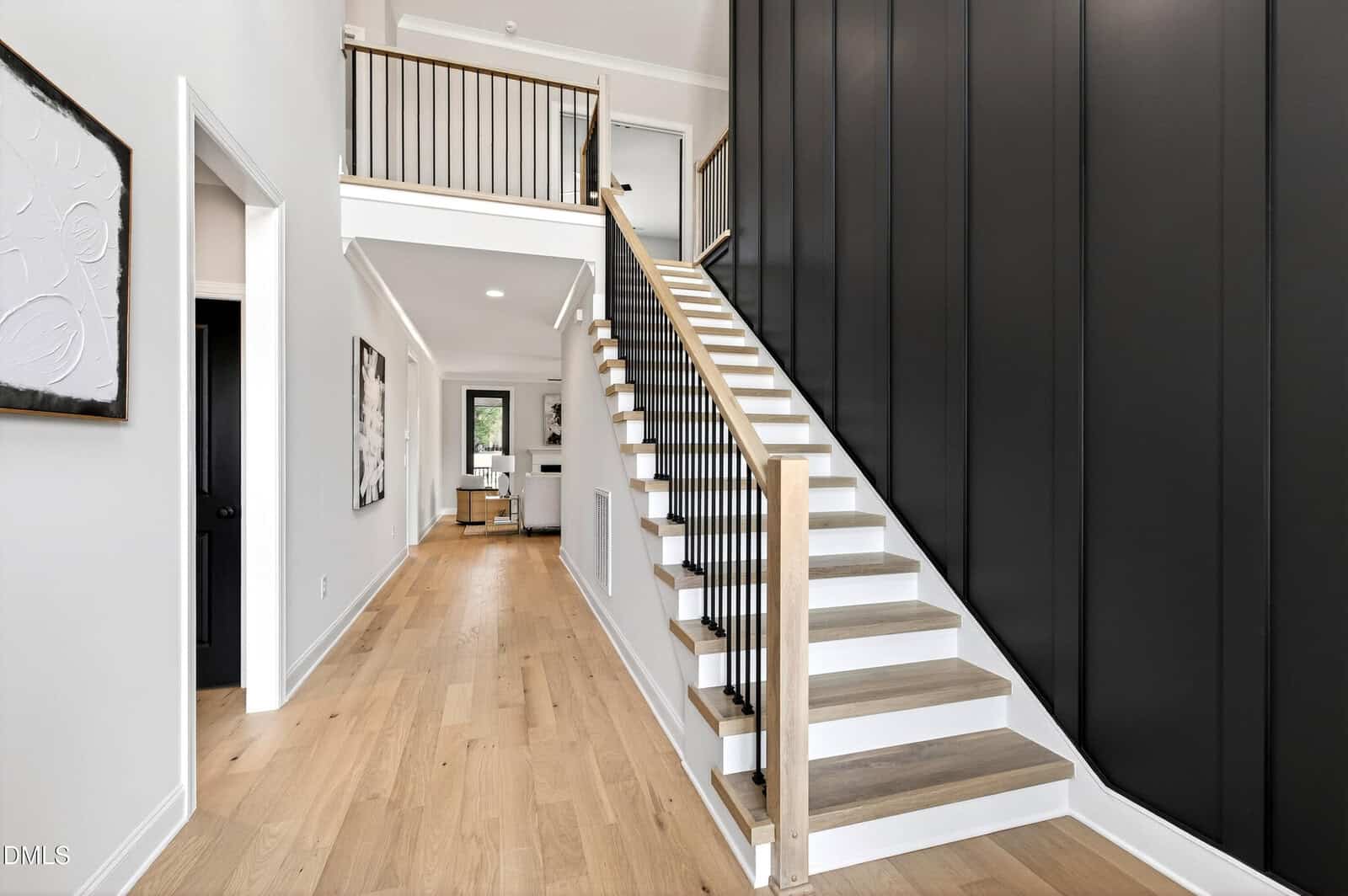 Modern home interior with light wood flooring, a staircase with wooden steps and black railings, and a tall matte black accent wall. Hallway leads to a bright living area with large windows. Minimalist decor and clean lines throughout.