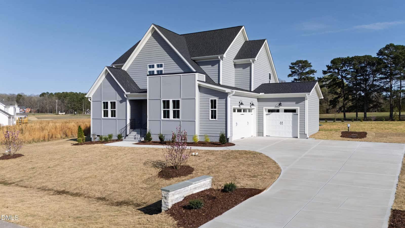Modern two-story gray house with a double garage, white trim, and a large concrete driveway, set on a spacious lot with minimal landscaping and surrounded by open fields and tall trees under a clear blue sky.