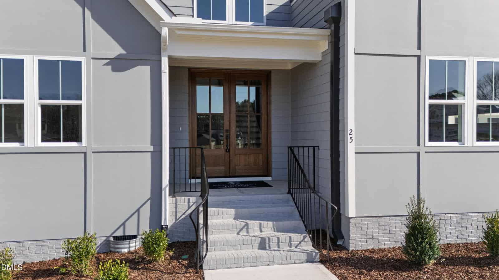 Front entrance of a modern house with light gray siding, large wooden double doors, white trim, several windows, black metal railings, concrete steps, small green shrubs, and a welcome mat.