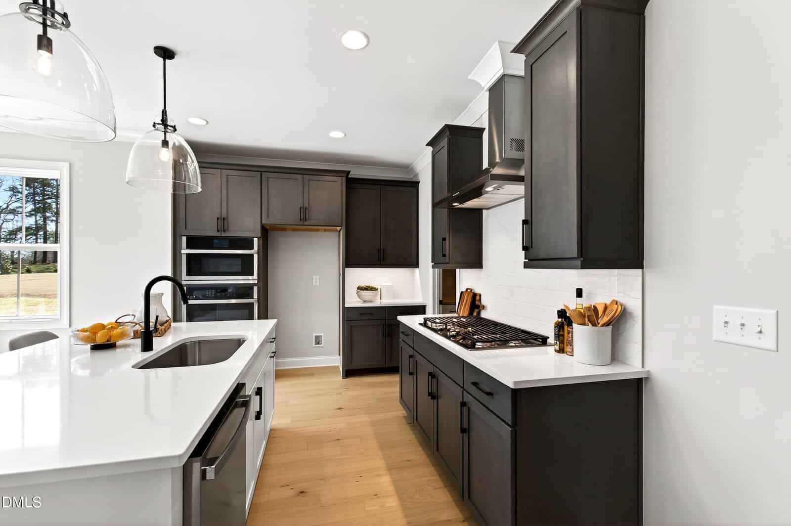 Modern kitchen with dark cabinets, white countertops, stainless steel appliances, a gas cooktop, island sink, pendant lights, and wooden floors. A large window lets in natural light.