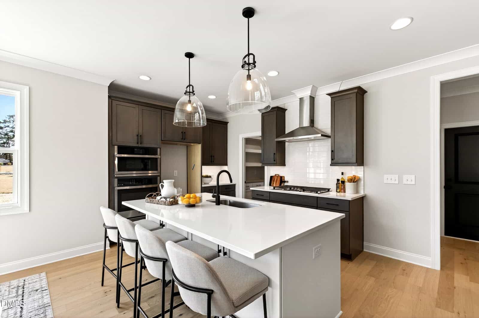 Modern kitchen with dark cabinets, white countertops, a central island with four upholstered stools, pendant lights, a built-in oven, gas cooktop, and wooden floors. A window lets in natural light.
