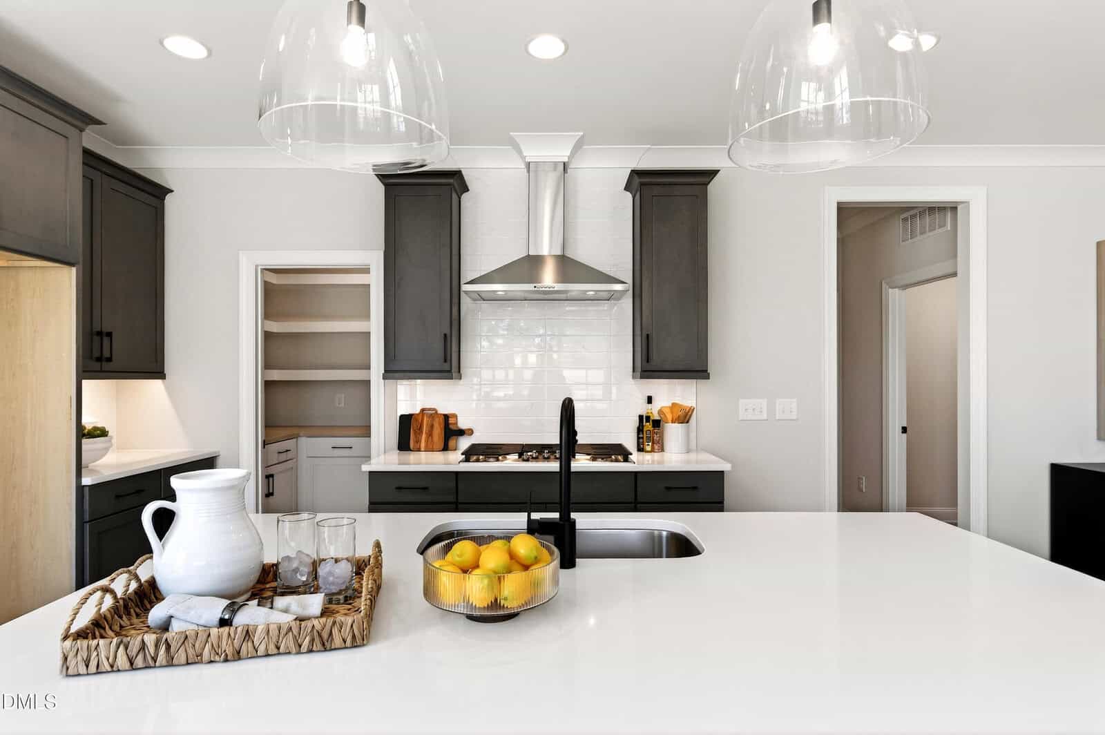 Modern kitchen with dark cabinets, white countertops, a central island with a sink, a tray with a pitcher and glasses, and a bowl of lemons. Stainless steel appliances and pendant lights are visible.