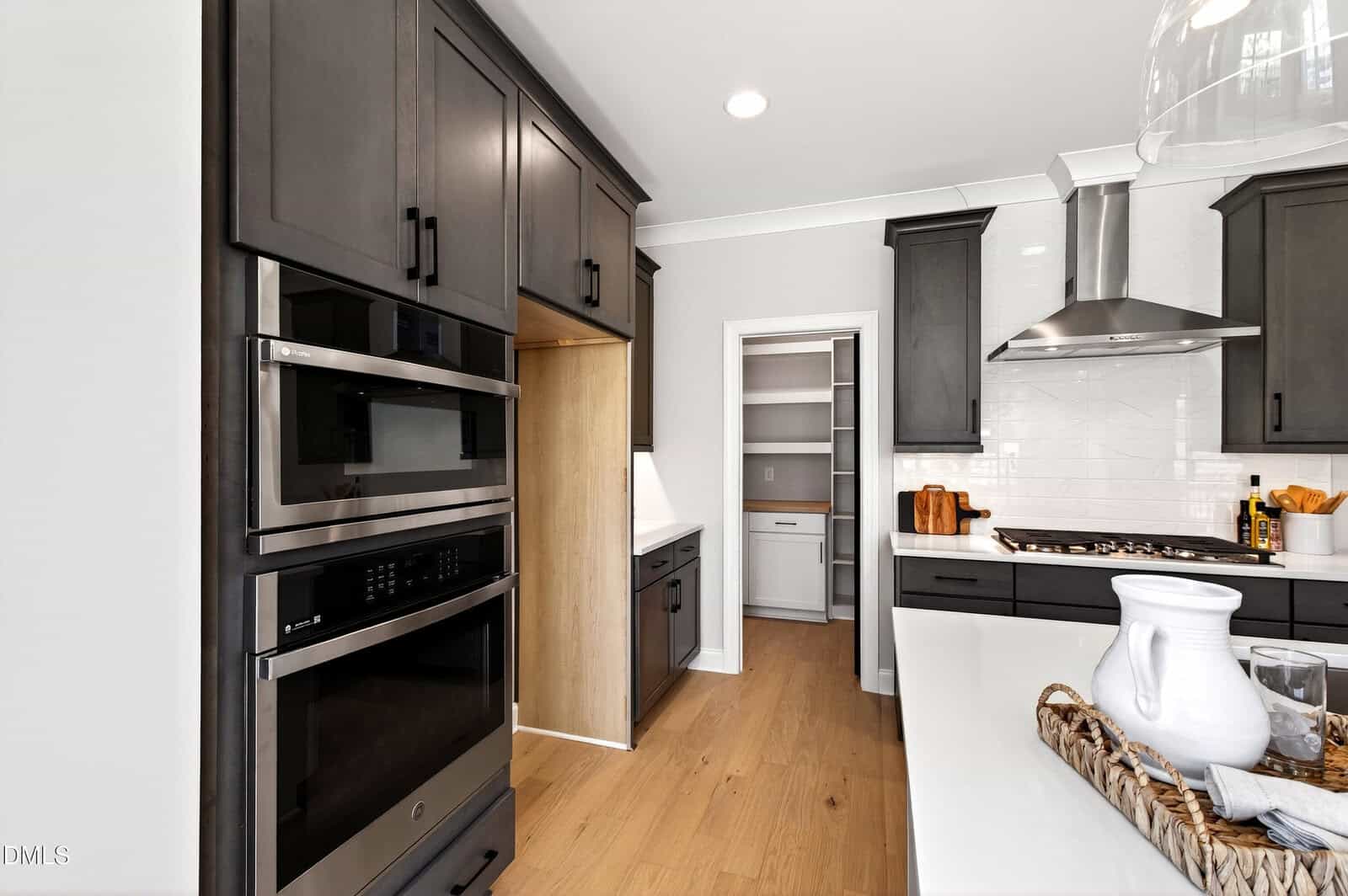 Modern kitchen with dark cabinets, double wall ovens, gas stove with hood, white countertops, wood floor, and walk-in pantry in the background. Countertop has a tray with a pitcher and cups.