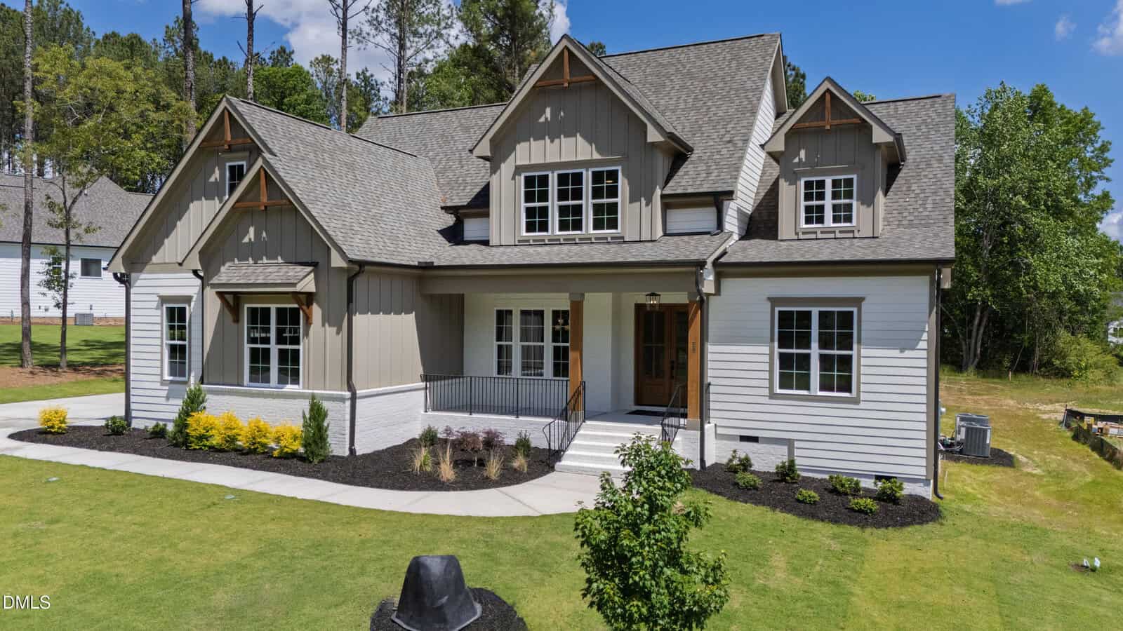 A modern two-story house with beige and white siding, large windows, landscaped front yard, and a covered front porch. Lush green lawn and trees surround the home on a sunny day.