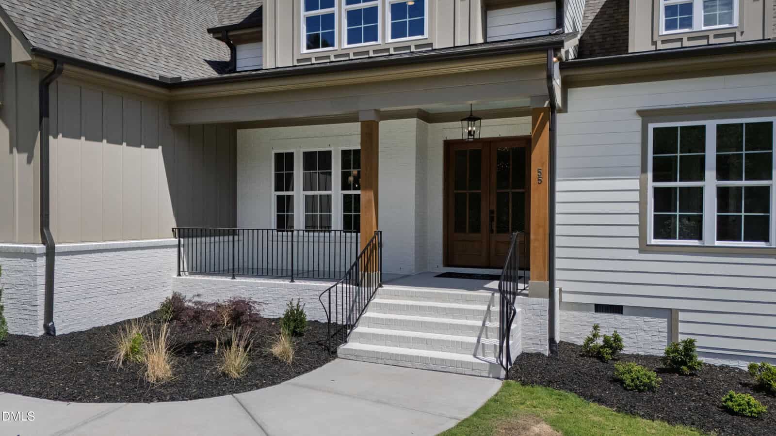 Modern house exterior with a covered front porch, wooden double doors, white brick walls, black metal railing, landscaped garden beds, and large windows. Concrete walkway leads to the entrance.