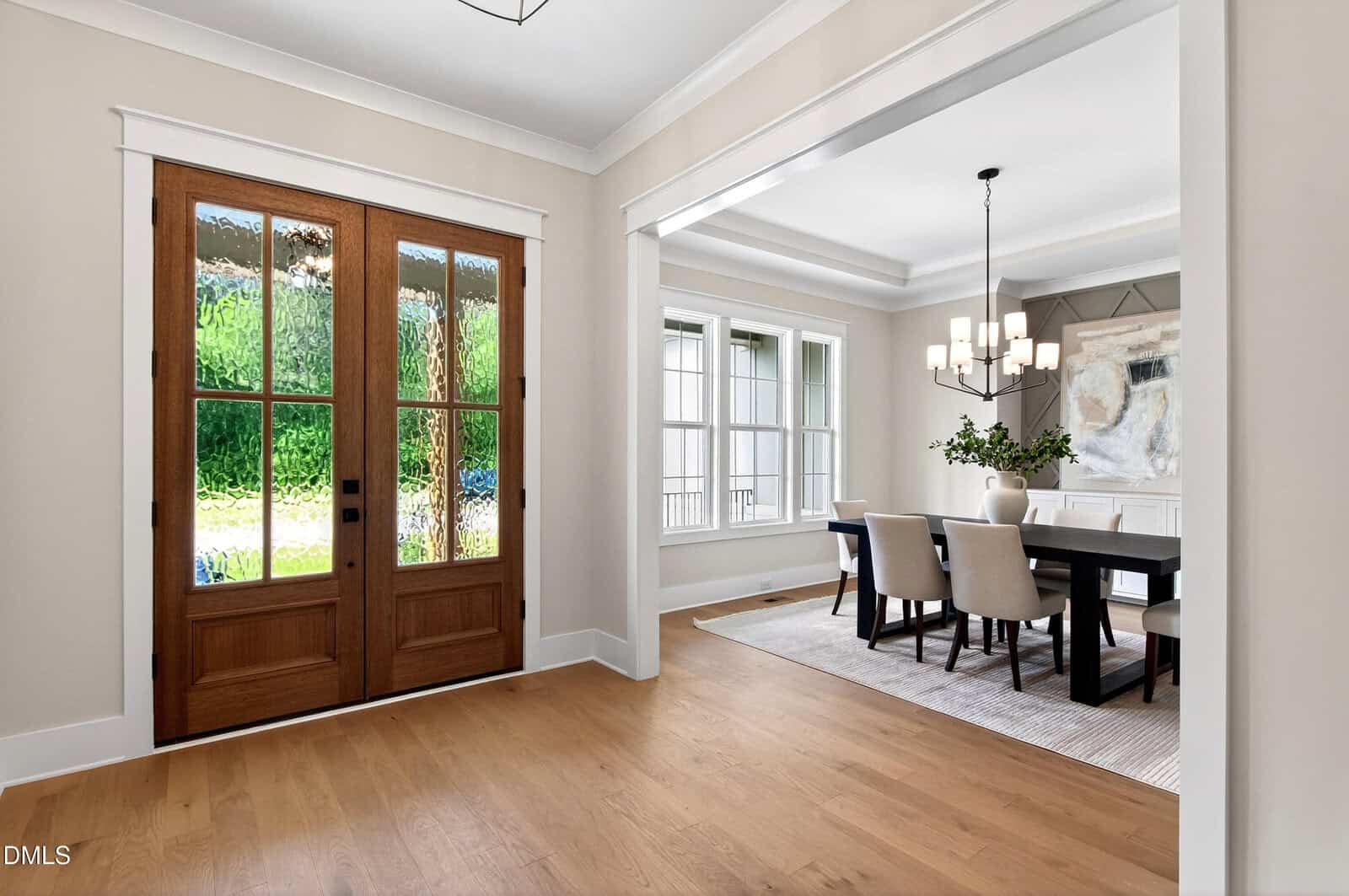 Bright entryway with double wooden doors, sidelights, and a view into a dining room featuring a dark table, six beige chairs, modern chandelier, large windows, and a neutral-toned painting on the wall.