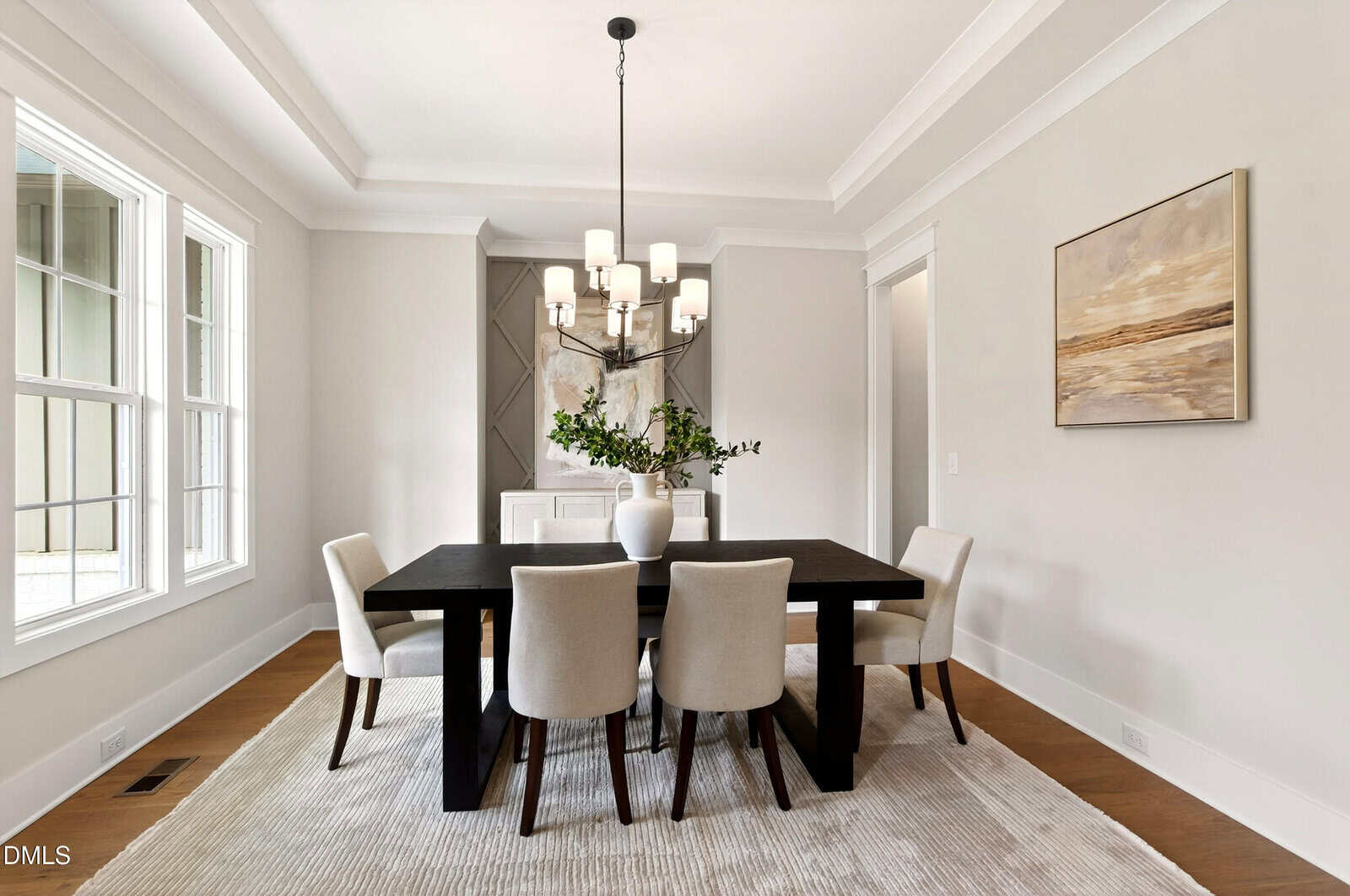 A modern dining room with a dark wooden table, six cream upholstered chairs, a chandelier above, large windows, a light area rug, a sideboard, and abstract artwork on the wall.