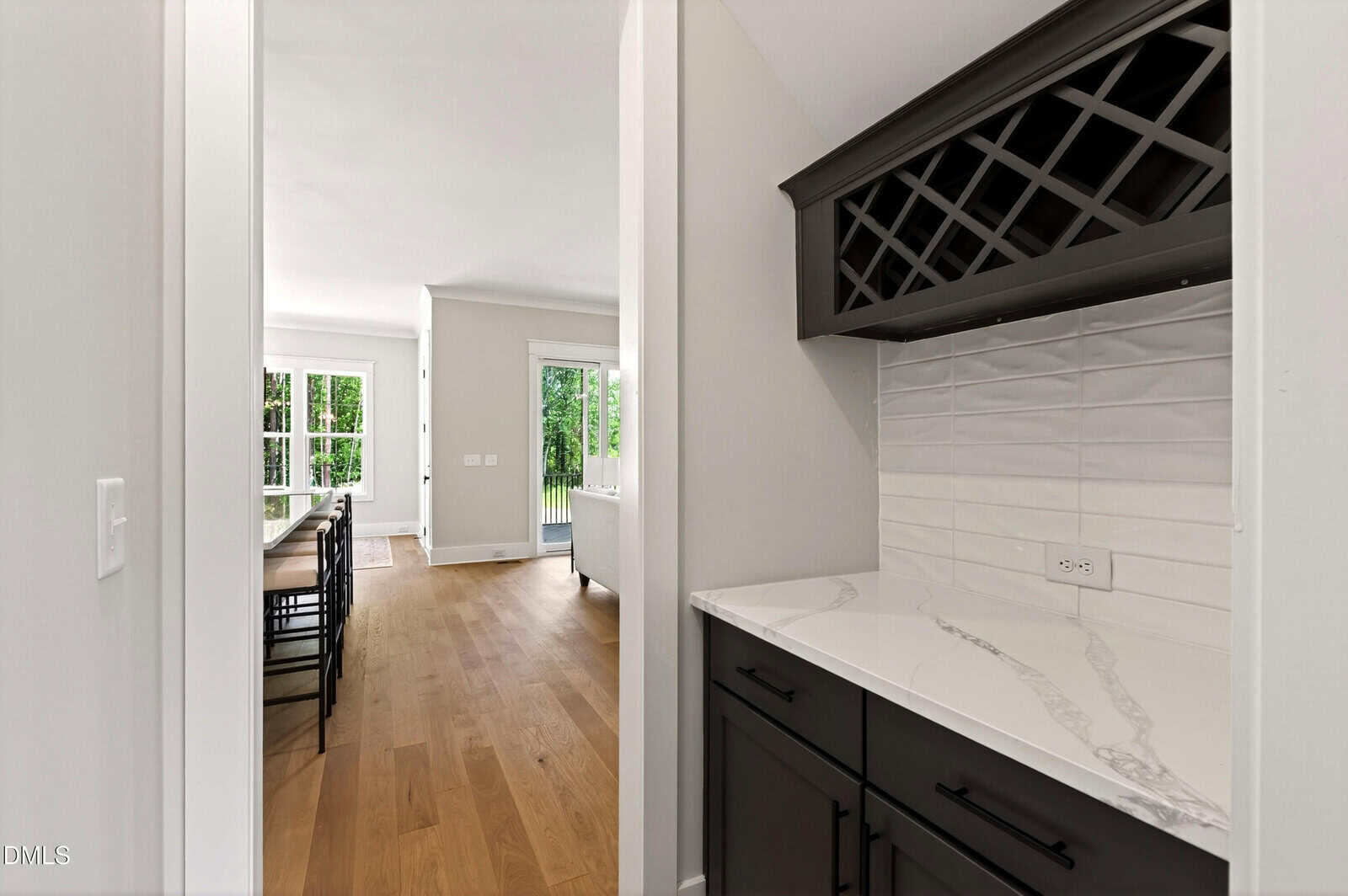 Modern kitchen nook with black cabinets, a white marble countertop, a built-in wine rack, and white tile backsplash. Wooden floors lead to a bright dining area with large windows and a door to the outside.