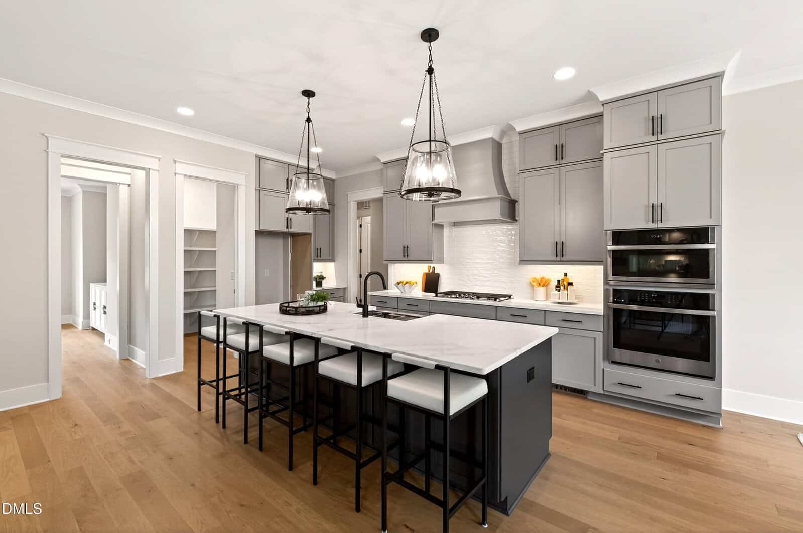 Modern kitchen with grey cabinets, stainless steel appliances, a white tile backsplash, large island with white countertop, black stools, pendant lights, and wood flooring. Doorway leads to a pantry and another room.