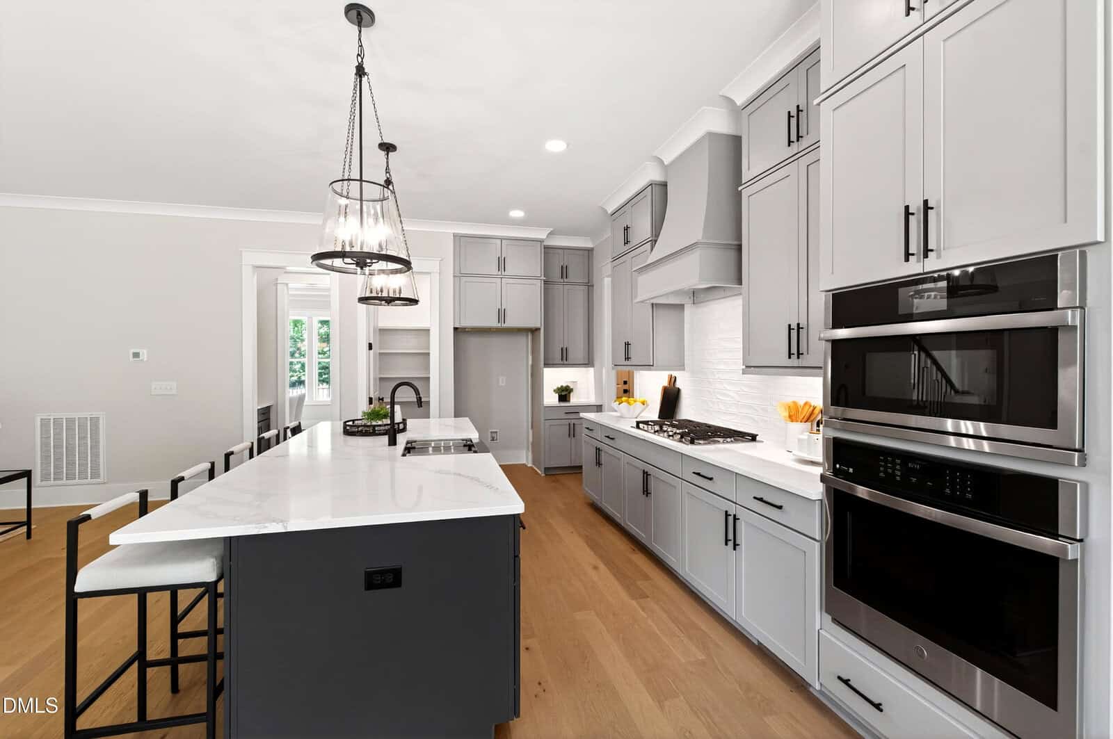 Modern kitchen with gray cabinets, white countertops, stainless steel appliances, gas stovetop, pendant lights, and light wood floors. There is a large island with bar stools and windows in the background.