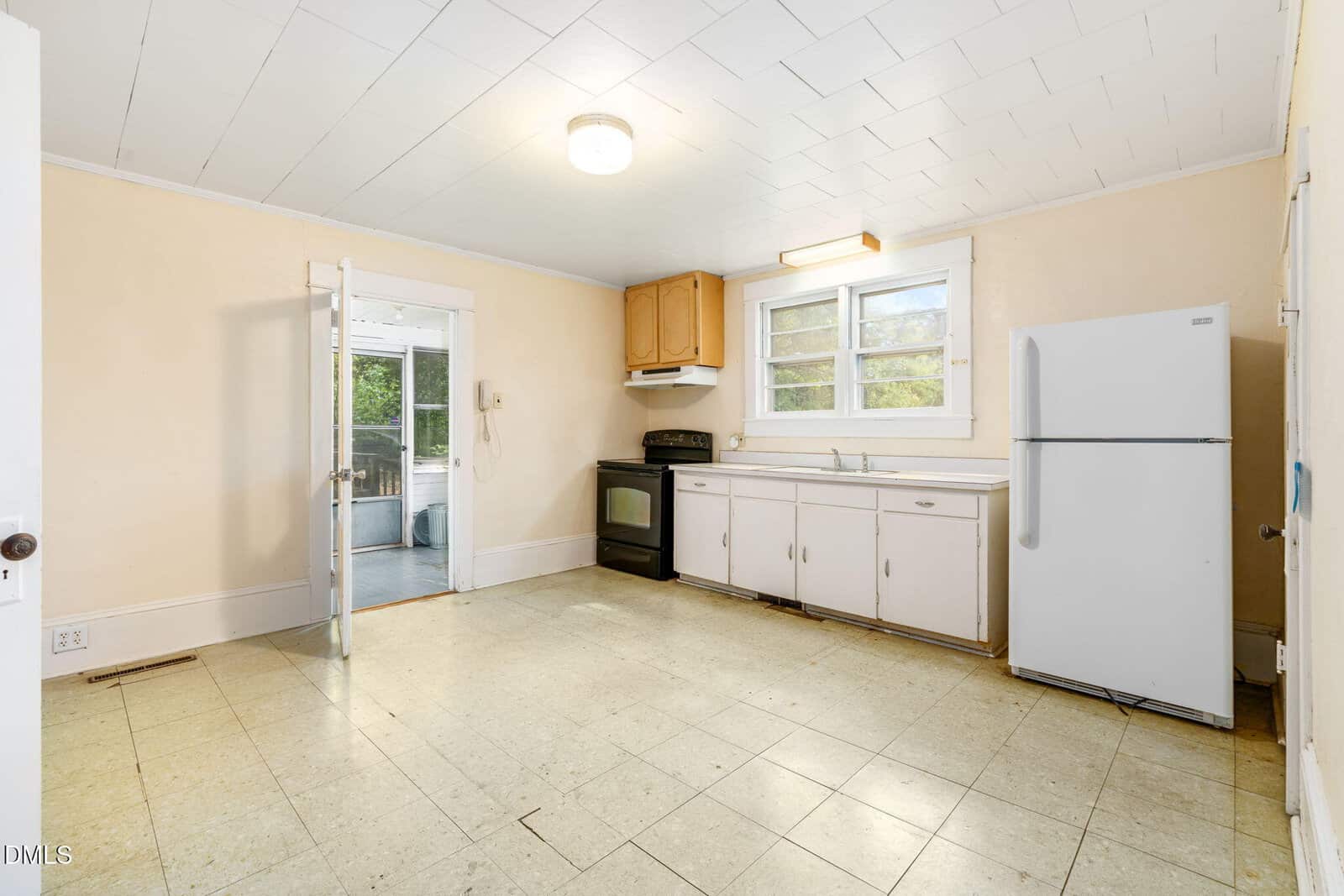 An empty kitchen with a white ceiling, light yellow walls, and a linoleum floor. The room has a stove, white refrigerator, cabinets, a window over the sink, and a door leading to an adjacent sunroom.