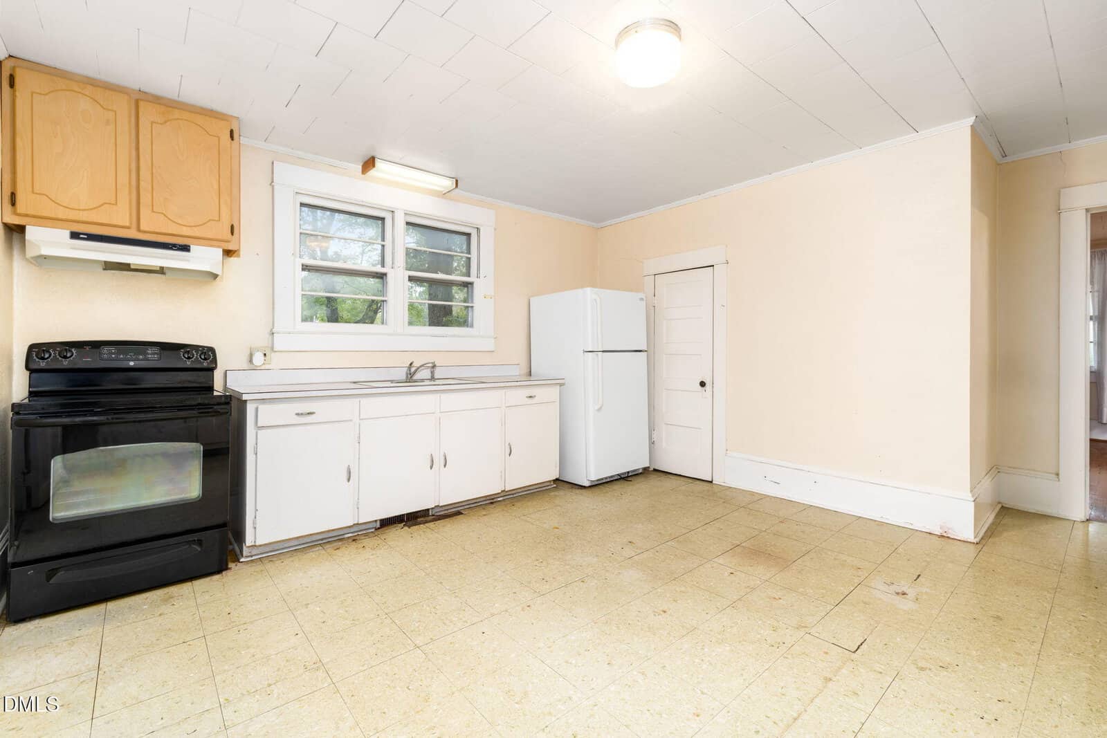 A simple kitchen with a black stove, white cabinets, a white refrigerator, and a small white door. There are two windows above the sink and beige walls with a tile floor.