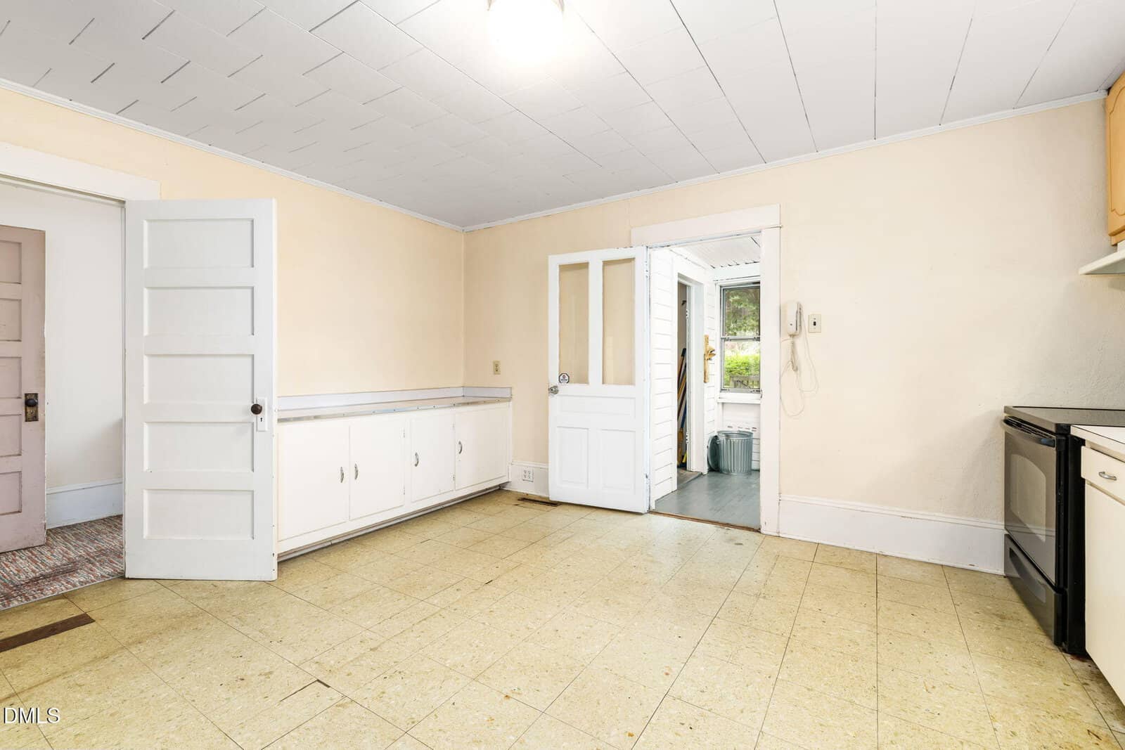 A spacious, empty kitchen with cream walls, a tiled ceiling, linoleum floors, white cabinets along one wall, an open door leading to another room, and a glass door to a sunroom or porch.