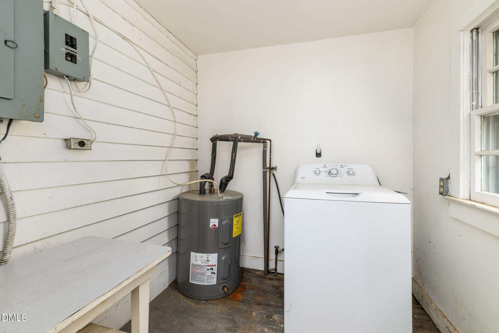 A small laundry room with a white top-load washing machine, a gray water heater, a wall-mounted fuse box, white wood-paneled walls, a table, and a window letting in natural light.
