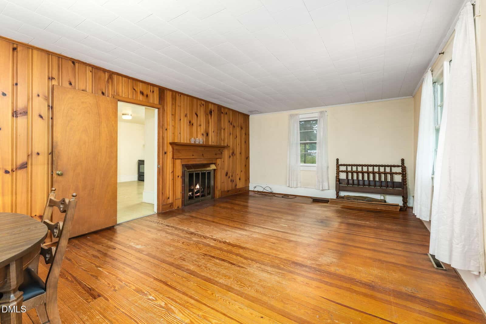 A living room with wood-paneled and cream-colored walls, hardwood floor, a fireplace, two windows with white curtains, a wooden bench, and part of a round dining table and chairs in the foreground.