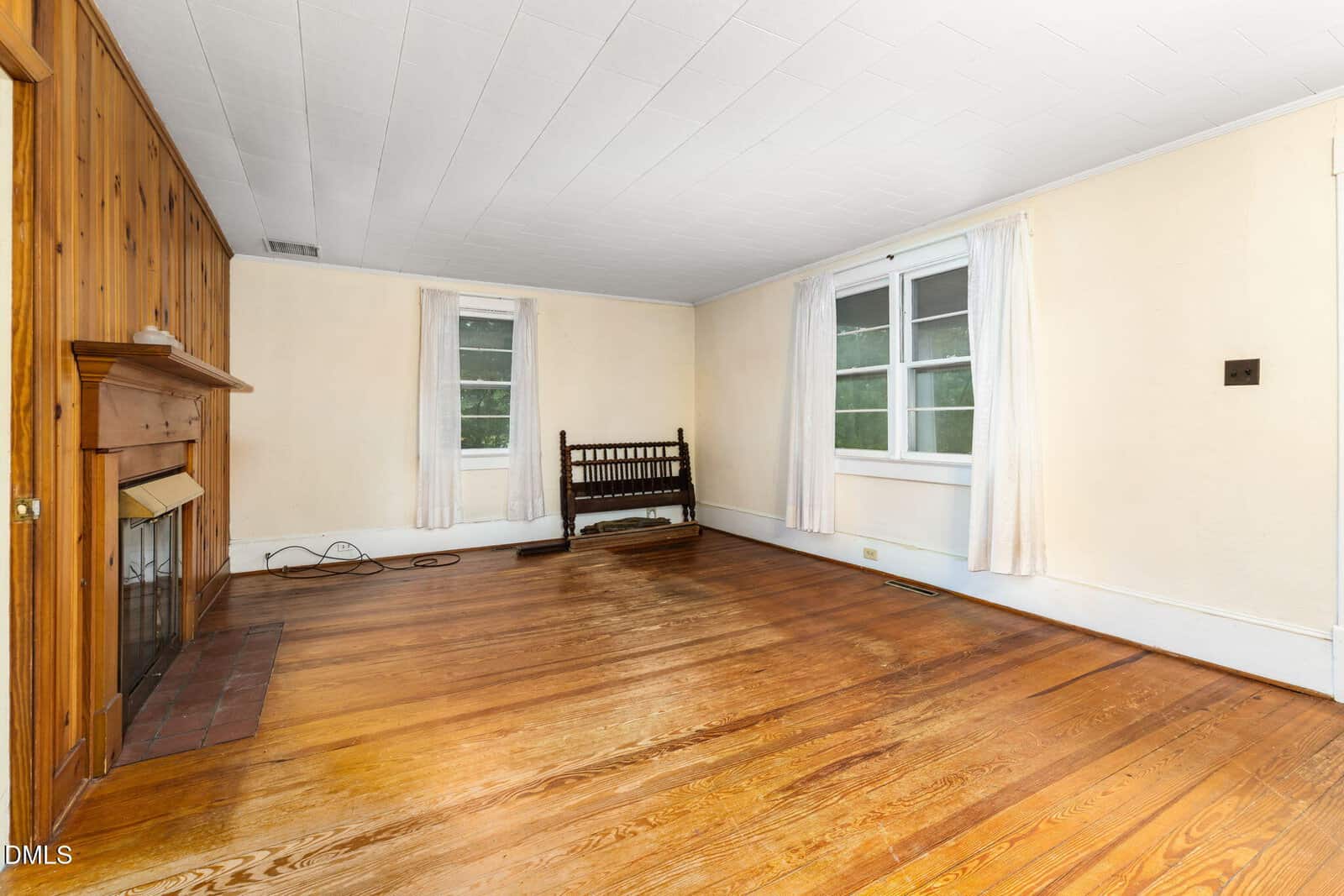 A mostly empty living room with wood floors, a wooden fireplace on the left wall, two windows with white curtains, cream-colored walls, and a wooden bench along the back wall.