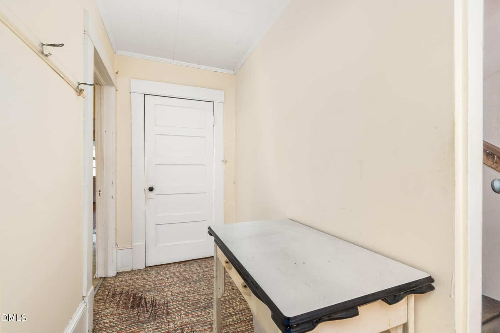 Narrow room with beige walls, a white door at the far end, a small table with a white top and black edges, worn carpet, and a staircase partially visible to the right.
