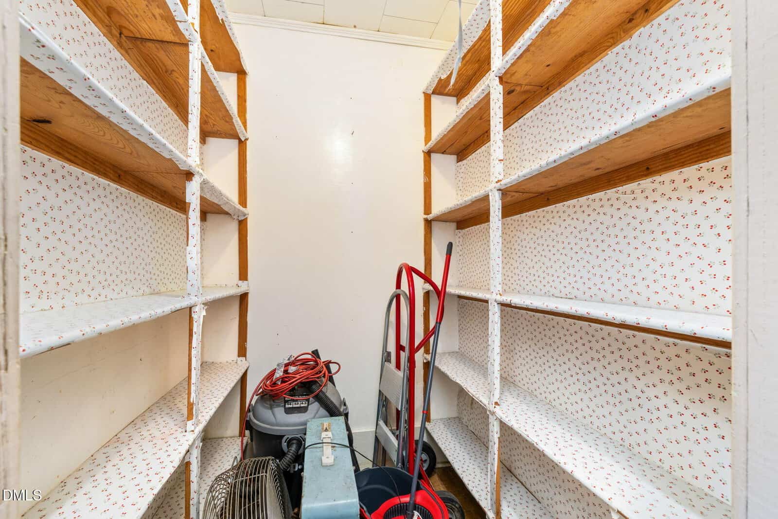 A small storage room with shelves on both sides, lined with patterned paper. Cleaning equipment and supplies, including a vacuum, fan, and mop bucket, are placed on the floor at the back of the room.