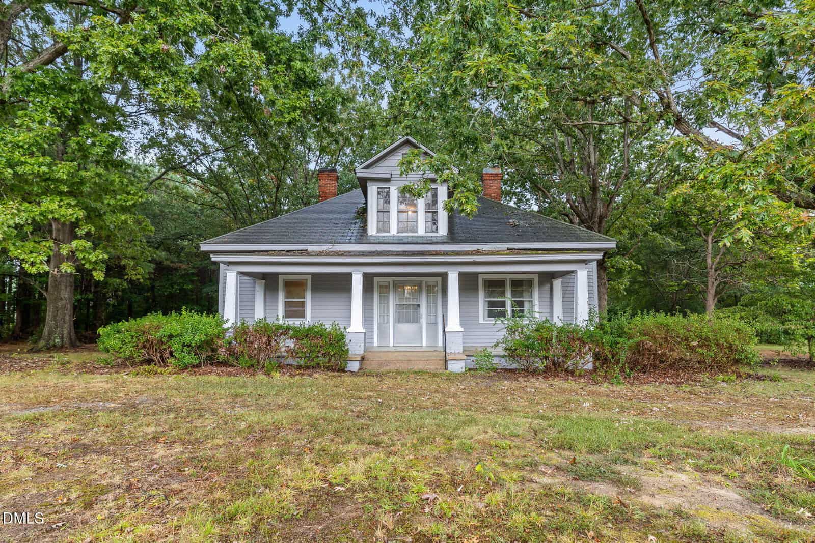 A small, light gray house at 1089 Gillburg Road in Henderson features a covered front porch, white trim, and a dormer window. It sits among tall trees and bushes with a patchy dirt and grass yard in front.