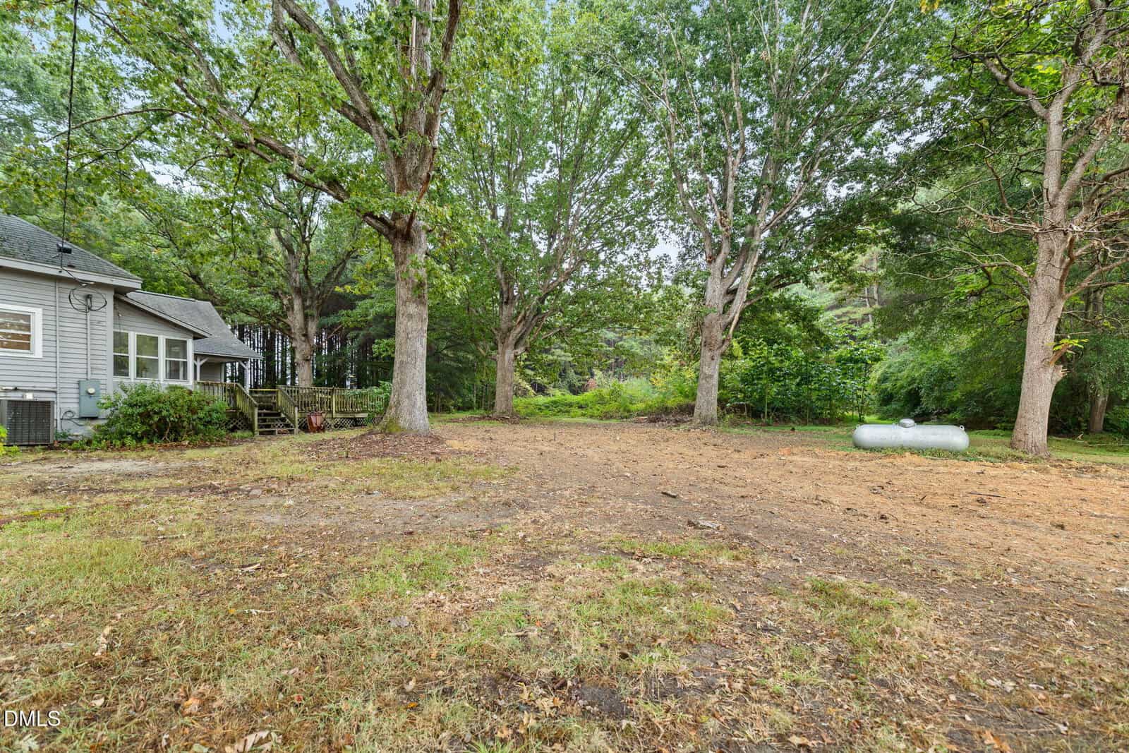 A backyard with large mature trees, a mostly bare and patchy lawn, a propane tank on the right, and part of a gray house with a wooden deck on the left. The area is bordered by dense greenery.