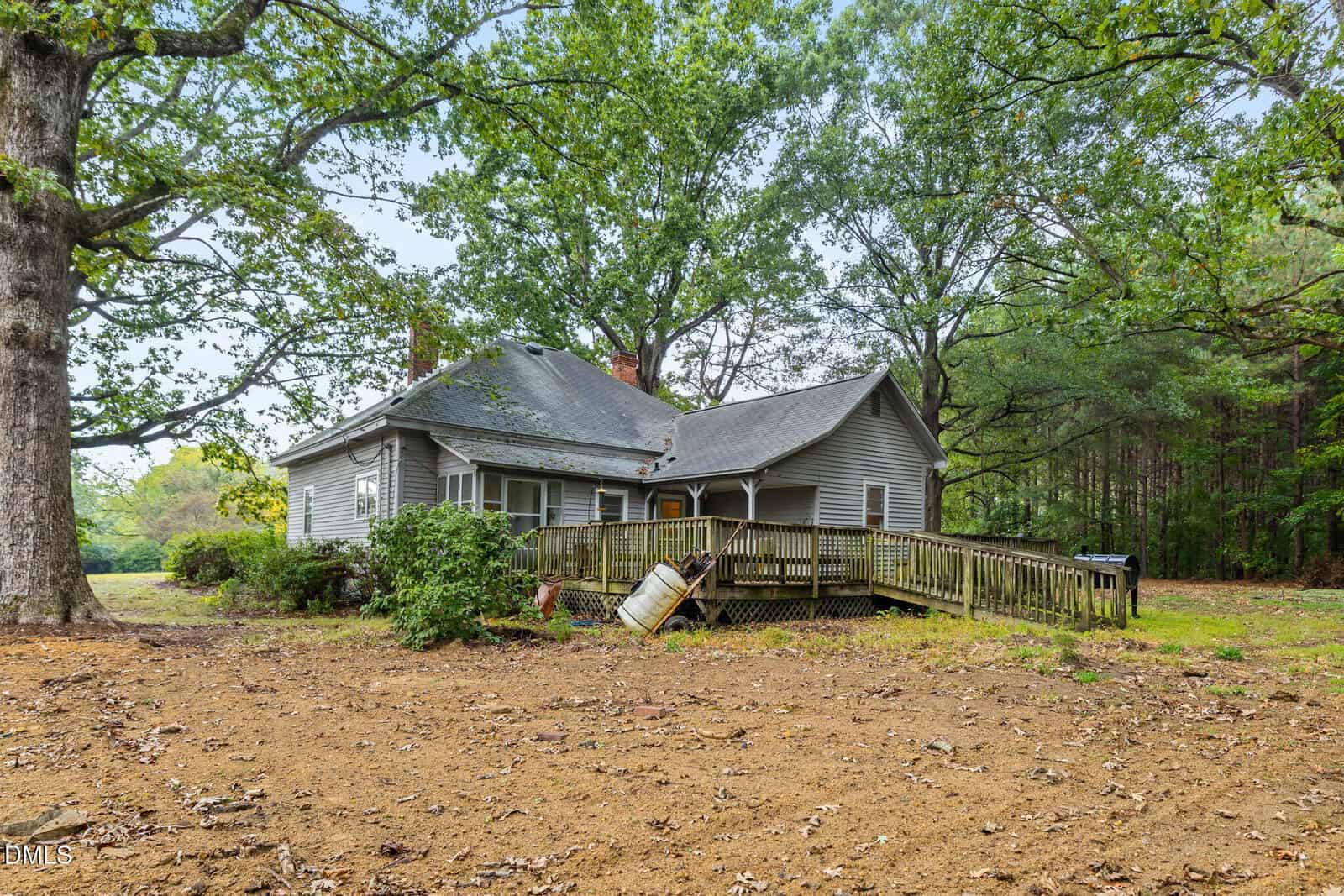 A gray, single-story house with a sloped roof, brick chimneys, and a wooden ramp leading to the porch. The house is surrounded by large trees, sparse grass, and a mostly bare dirt yard.